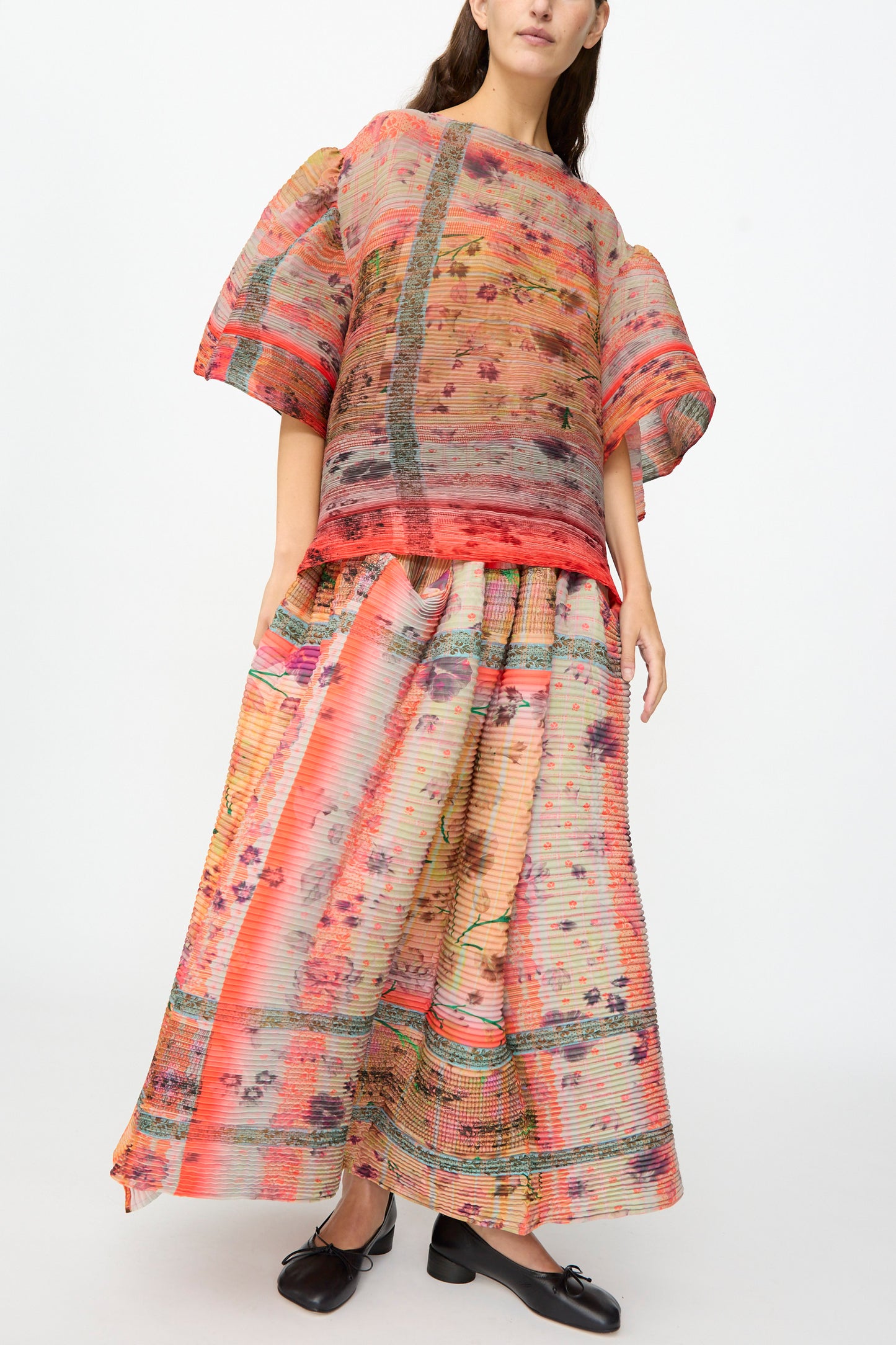 A woman stands against a plain light background wearing the Julia Heuer Robin Handpleated Top in Bruno—a loose, colorful, patterned sculptural top with short sleeves crafted from recycled chiffon—paired with a matching wide skirt and black shoes.
