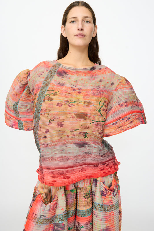 A woman stands against a plain white background, wearing the Julia Heuer Robin Handpleated Top in Bruno—a sheer, multicolored, pleated top with floral patterns and a subtly sculptural silhouette—paired with a matching recycled chiffon skirt.