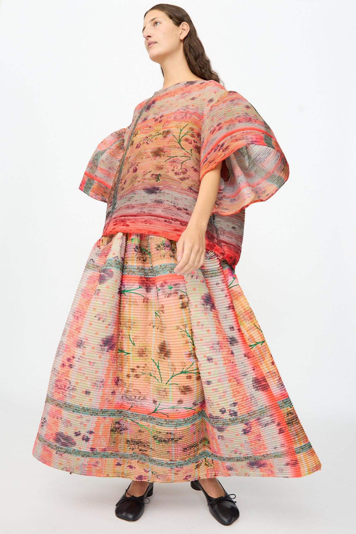 A person is wearing the Julia Heuer Robin Handpleated Top in Bruno—an oversized, colorful, patterned piece crafted from recycled chiffon—with a matching long skirt and short sleeves, standing against a plain white background.