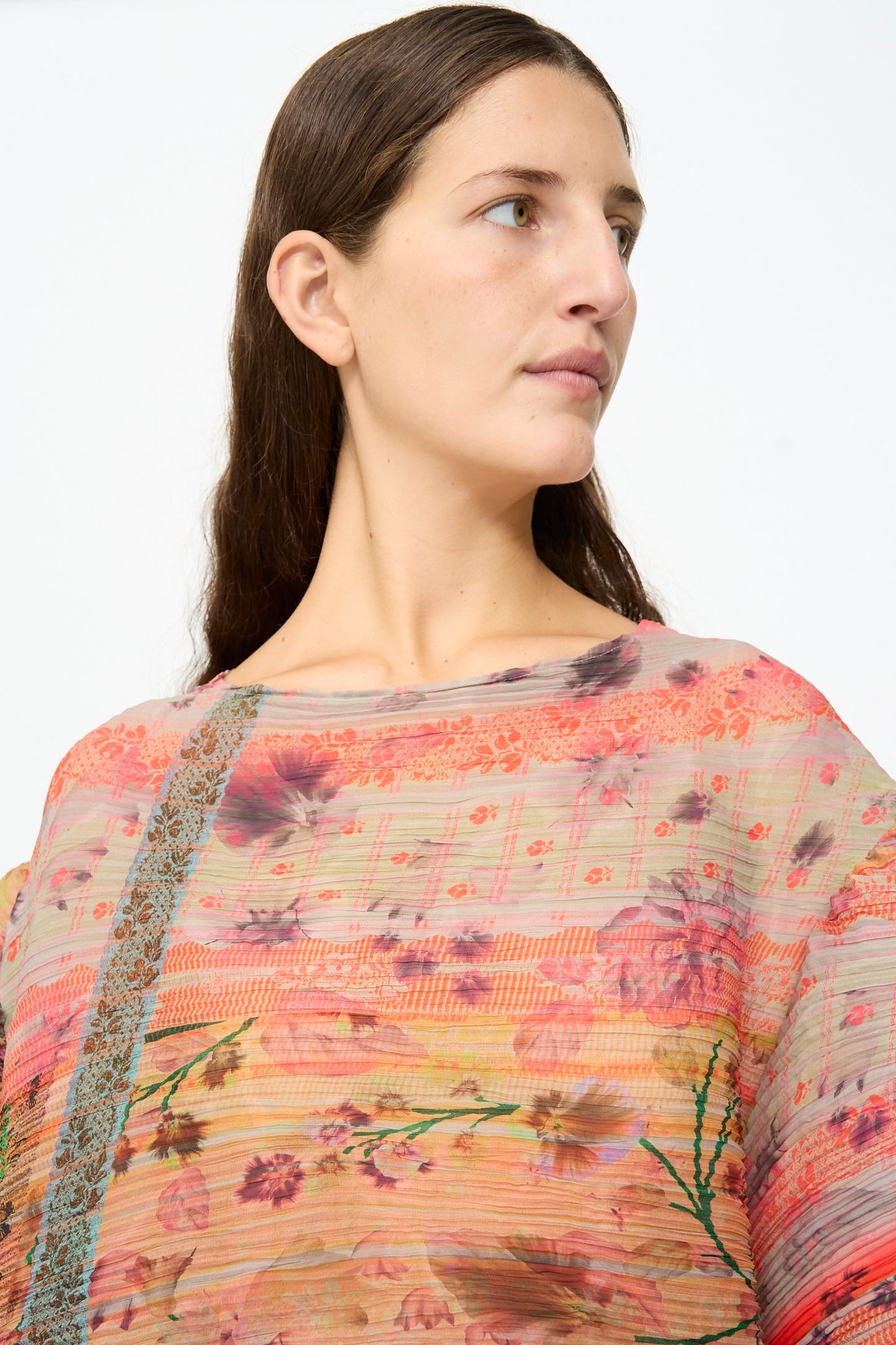A person with long brown hair wears the Julia Heuer Robin Handpleated Top in Bruno—a sheer, colorful floral-patterned piece crafted from recycled chiffon—and looks to the side against a plain background.