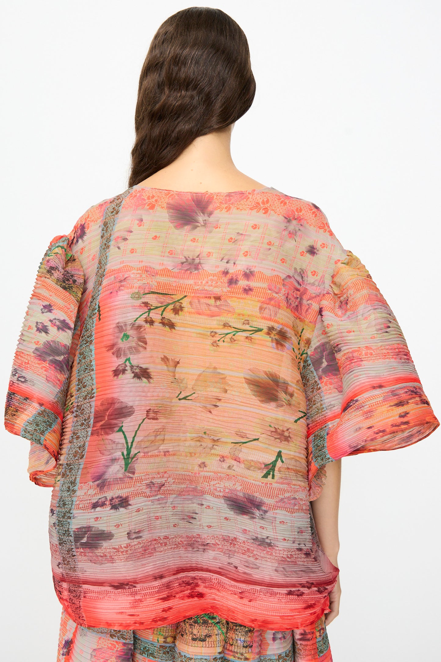 A person facing away, wearing the Julia Heuer Robin Handpleated Top in Bruno—an oversized, sheer, and loose-fitting sculptural piece crafted from recycled chiffon and adorned with floral and striped patterns in red, pink, and orange hues. Wavy brown hair drapes over one shoulder.