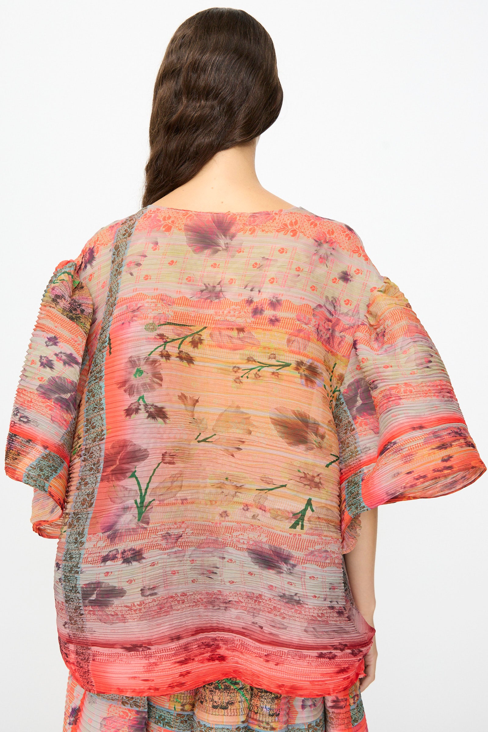 A person facing away, wearing the Julia Heuer Robin Handpleated Top in Bruno—an oversized, sheer, and loose-fitting sculptural piece crafted from recycled chiffon and adorned with floral and striped patterns in red, pink, and orange hues. Wavy brown hair drapes over one shoulder.