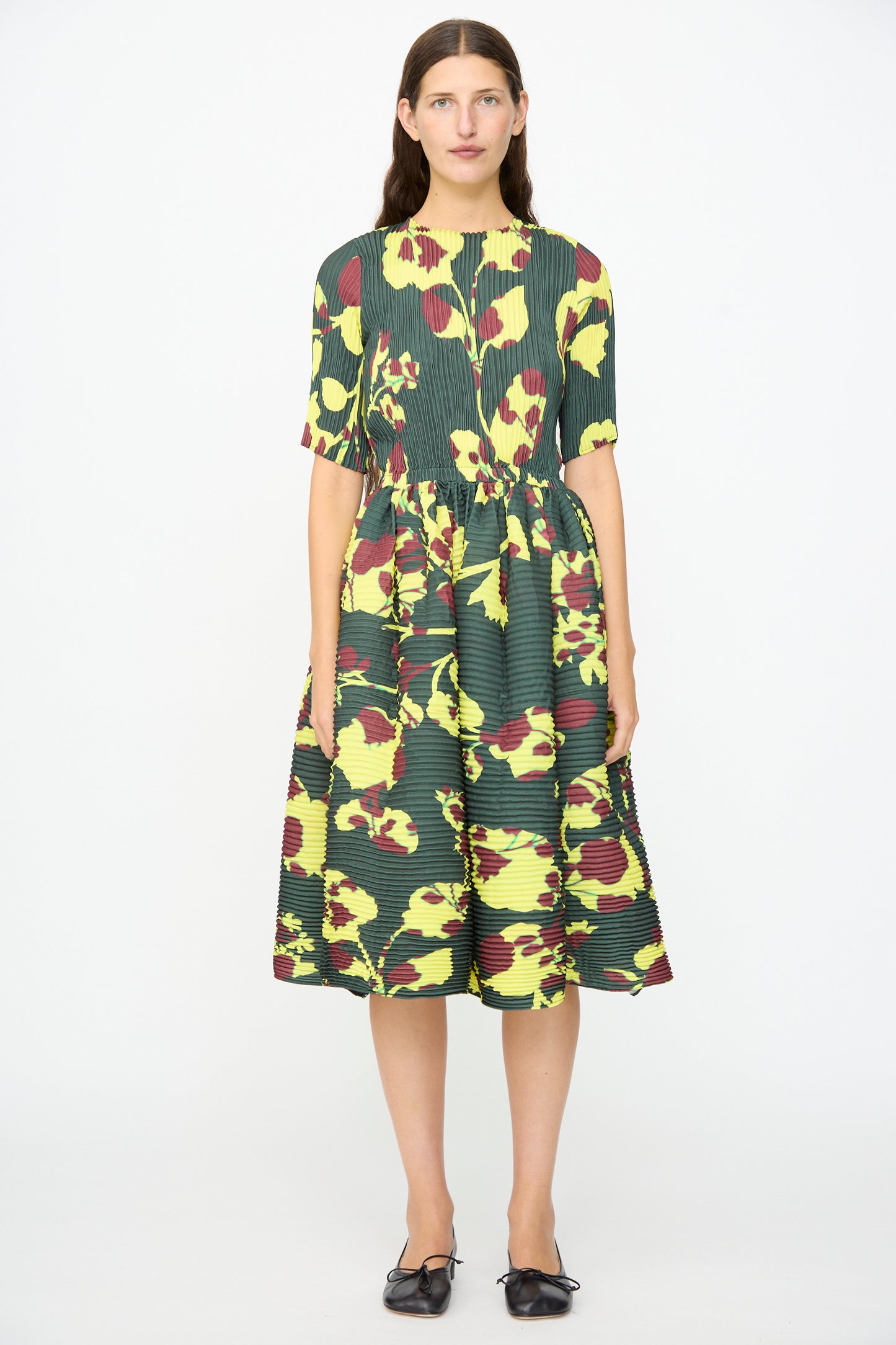 A woman stands facing forward wearing the limited-edition Julia Heuer Rocky Handpleated Dress in Electric Ingrid—a dark green, half-sleeve midi dress featuring yellow and red abstract floral patterns, crafted from recycled polyester crepe—paired with black shoes.