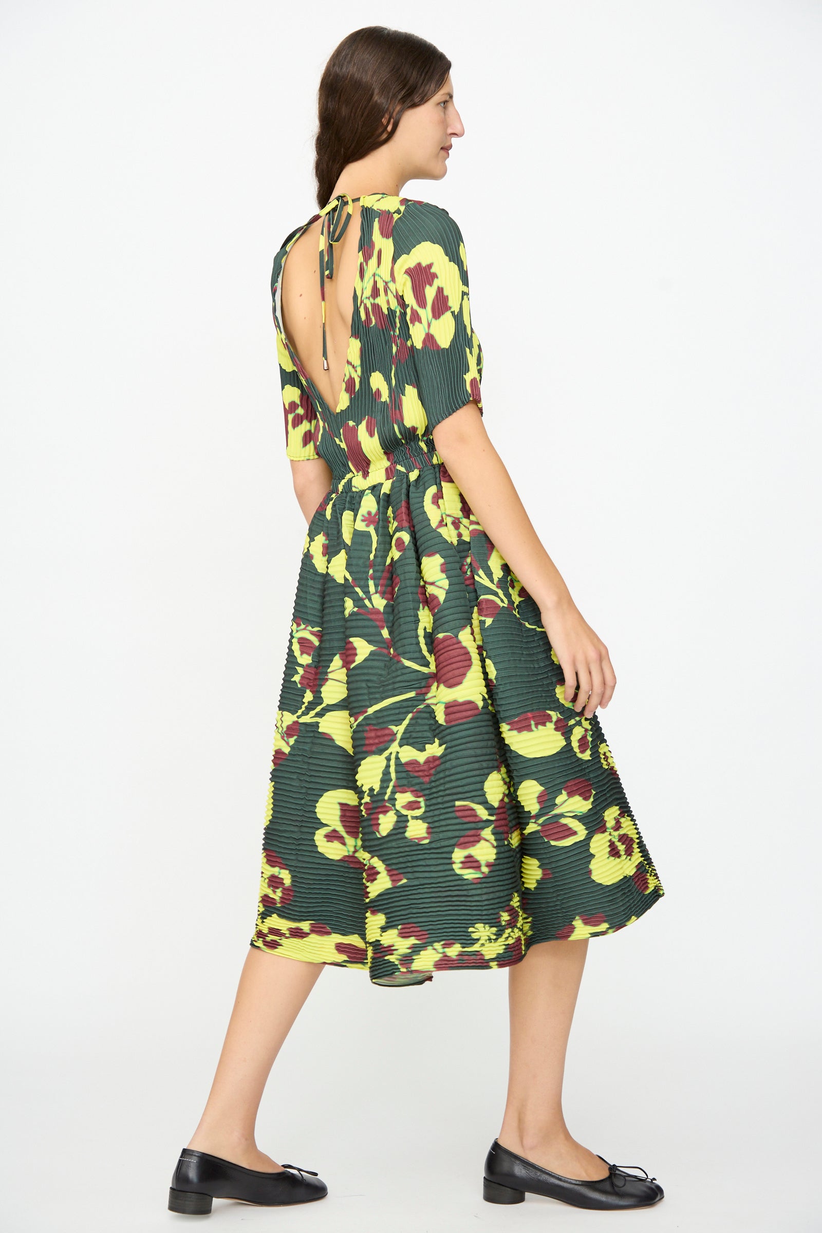 A woman wearing the Julia Heuer Rocky Handpleated Dress in Electric Ingrid—a limited edition, green floral half-sleeve midi dress made from recycled polyester crepe with an open back—pairs it with black flats as she stands against a plain white background, facing sideways.