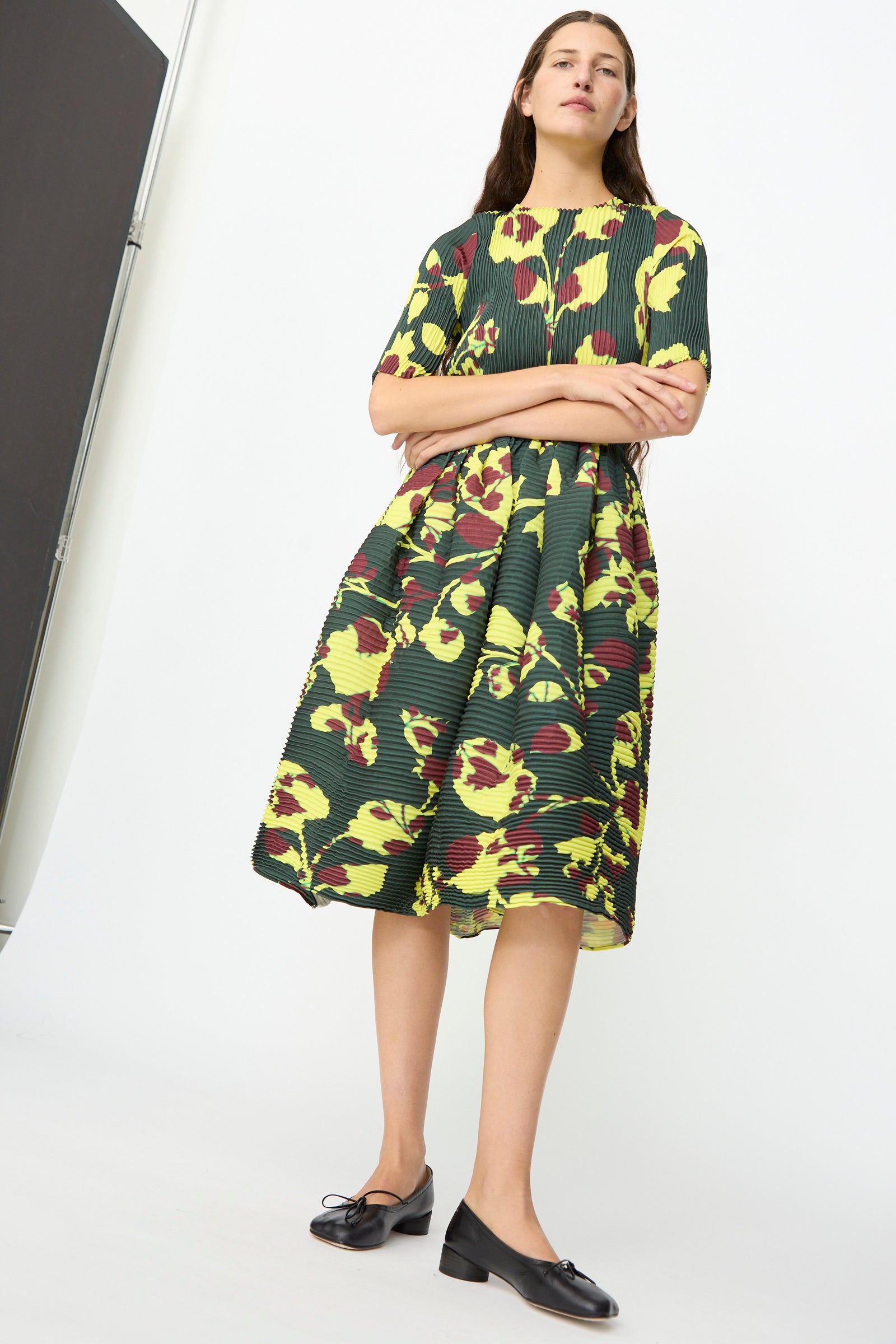 A woman stands with arms crossed, wearing the Julia Heuer Rocky Handpleated Dress in Electric Ingrid—a limited edition, half-sleeve midi dress crafted from recycled polyester crepe and featuring a vibrant green, yellow, and maroon floral pattern—paired with black loafers against a plain white background.