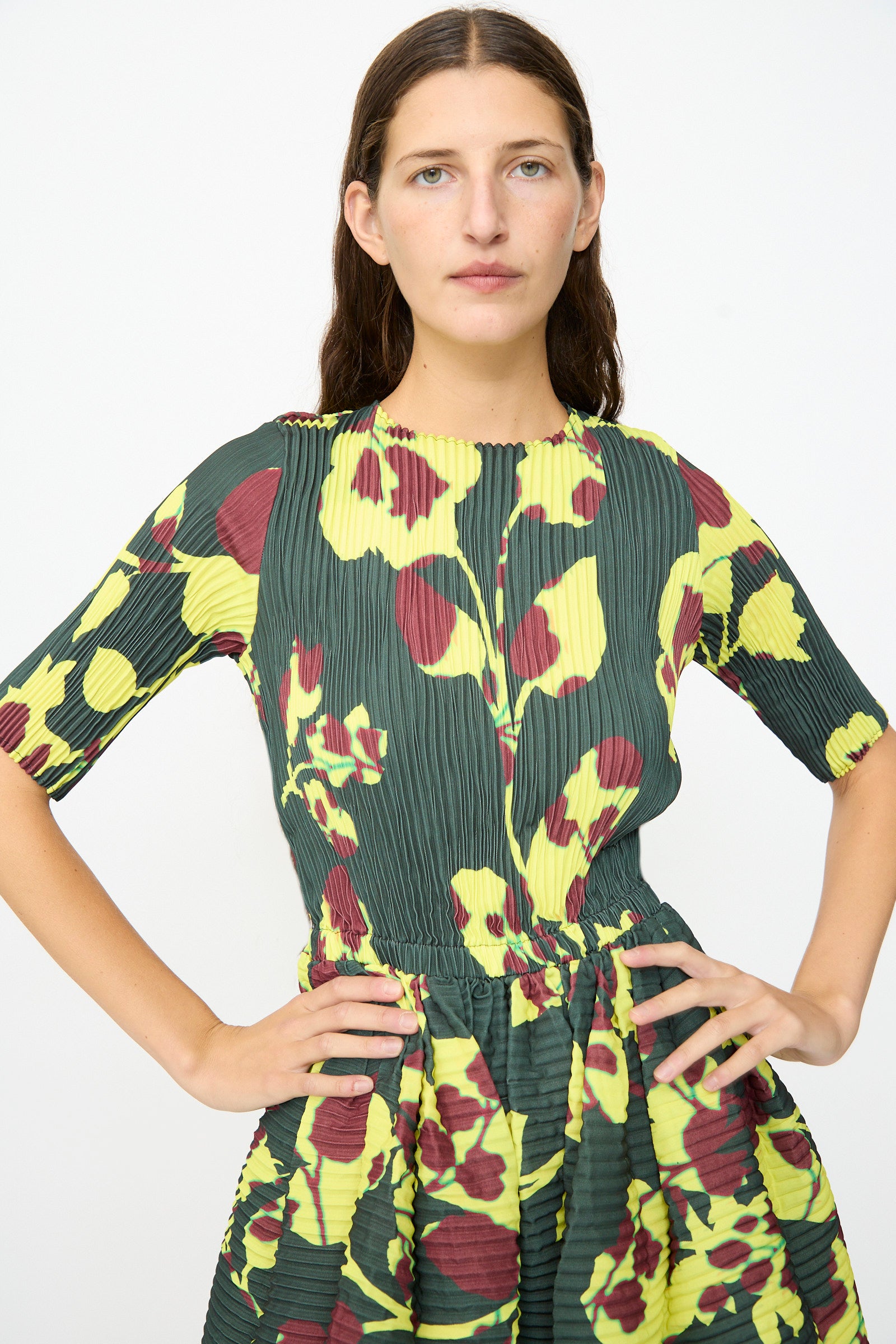 A woman stands with hands on hips against a plain white background, wearing the Julia Heuer Rocky Handpleated Dress in Electric Ingrid—a green half-sleeve midi dress featuring yellow and maroon floral patterns, expertly crafted from recycled polyester crepe.