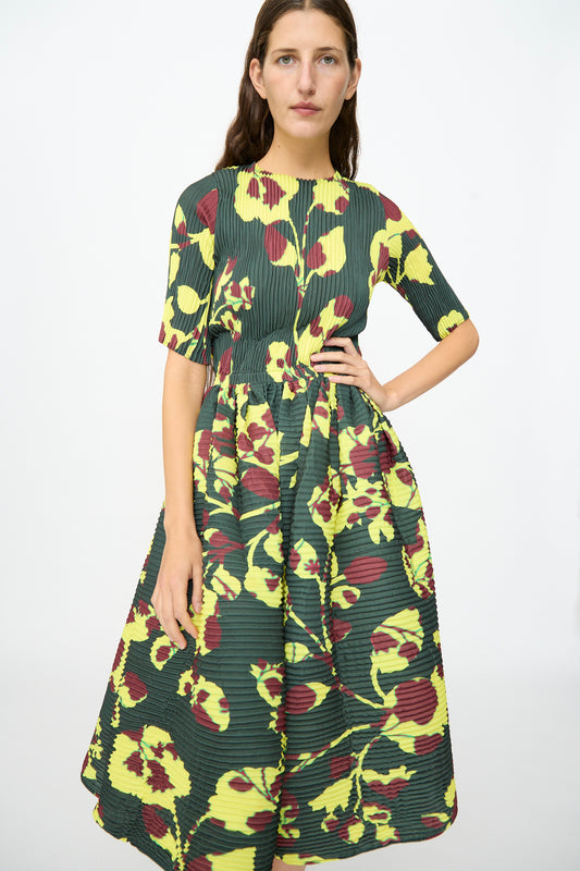A woman models the Julia Heuer Rocky Handpleated Dress in Electric Ingrid—a green, yellow, and maroon floral-patterned, pleated midi dress with short sleeves—crafted from recycled polyester crepe, standing against a plain white background.