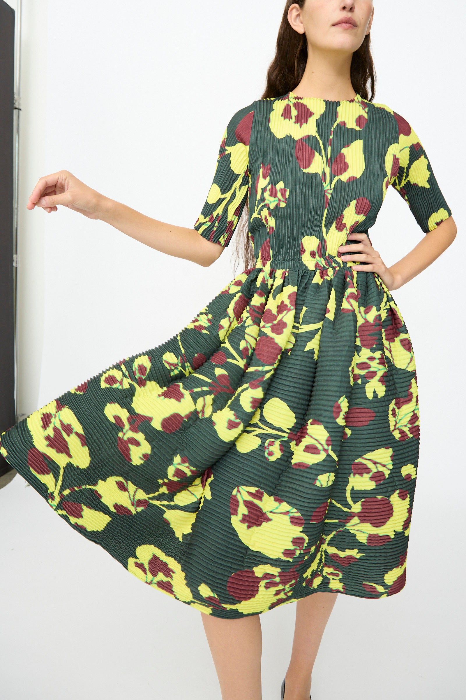 A woman poses with her hand on her waist and the skirt flared out, wearing the Julia Heuer Rocky Handpleated Dress in Electric Ingrid—a limited edition dark green midi dress with yellow and red floral print, crafted from recycled polyester crepe—against a plain background.
