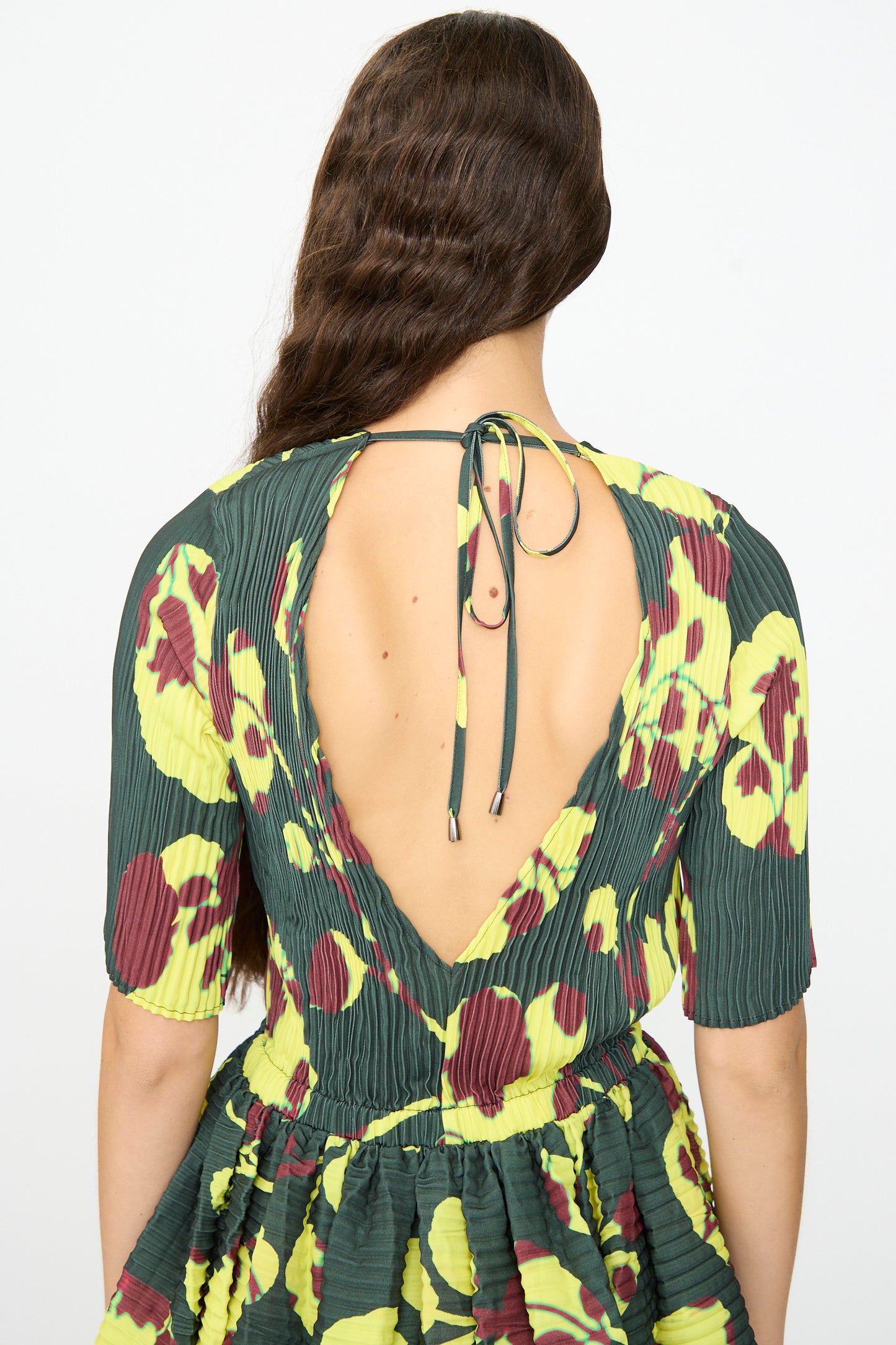 A woman with long, wavy brown hair wears the limited edition Julia Heuer Rocky Handpleated Dress in Electric Ingrid—a green half-sleeve midi dress crafted from recycled polyester crepe, decorated with a yellow and maroon floral pattern and featuring a deep V-back with a tie at the neck.