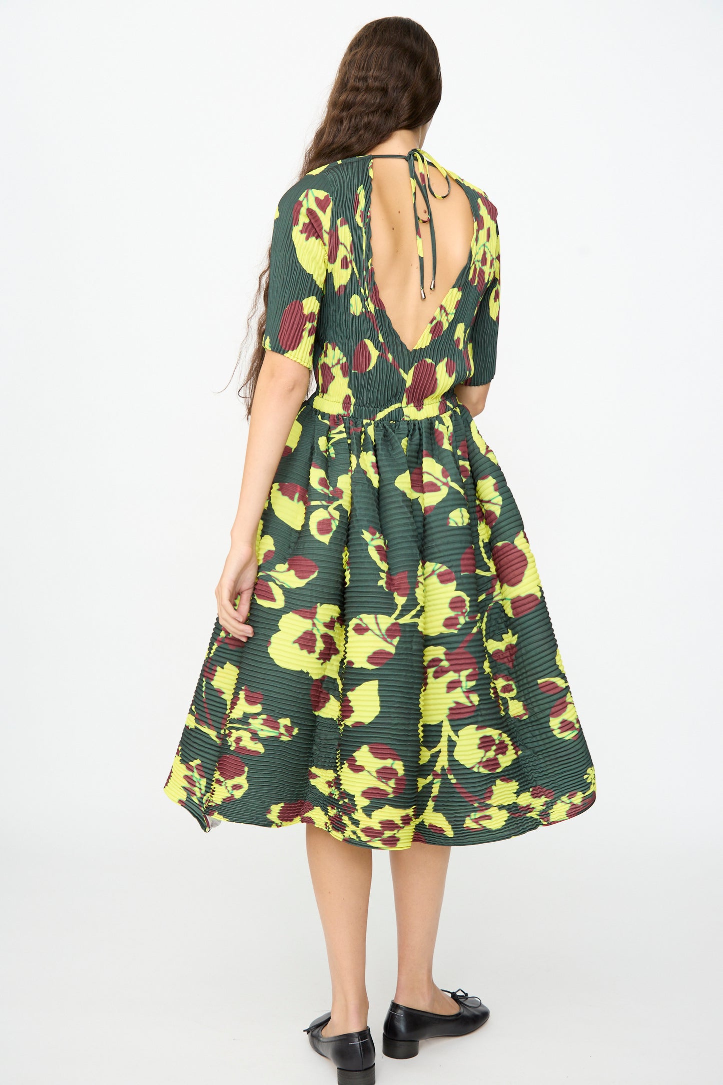 A woman with long hair wears the Julia Heuer Rocky Handpleated Dress in Electric Ingrid—a limited edition green and yellow floral half-sleeve midi dress crafted from recycled polyester crepe, featuring an open back—paired with black flats and standing against a plain white background.
