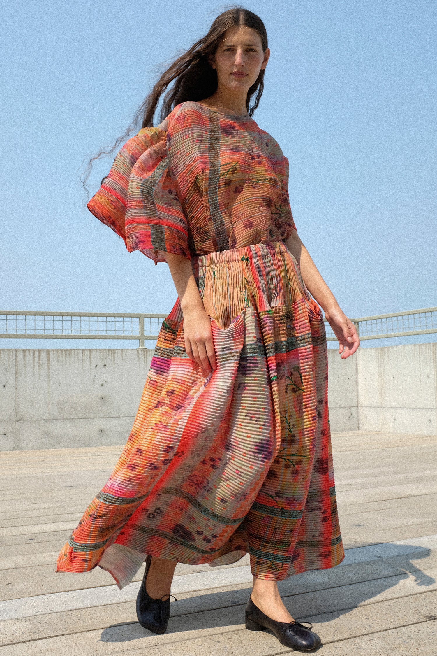 A woman wearing Julia Heuer's Robin Handpleated Top in Bruno, with Kenza Handpleated Skirt in Birte standing outdoors on a wooden deck under a clear blue sky.