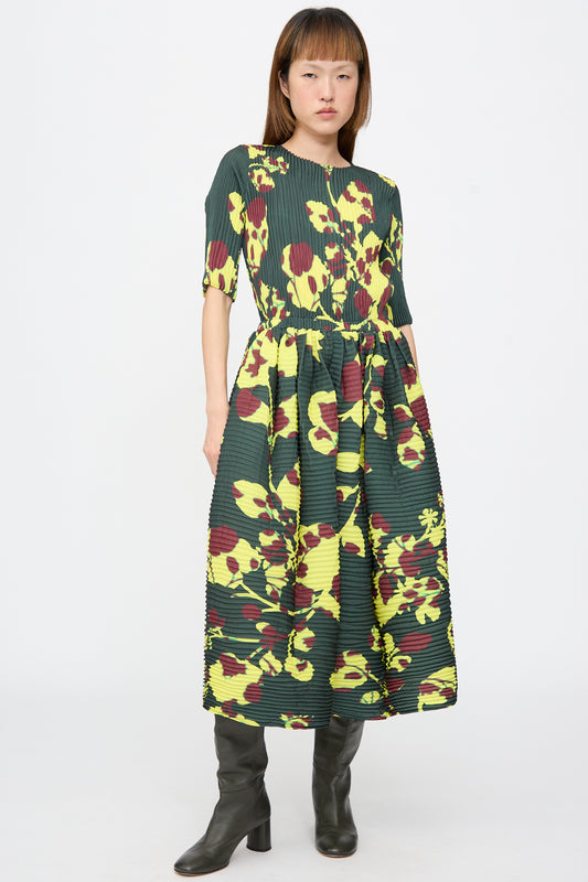 A model wears a Julia Heuer Rocky Handpleated Dress in Electric Ingrid, a dark green, hand-pleated crepe midi dress with a yellow and burgundy floral print, styled with black knee-high boots.