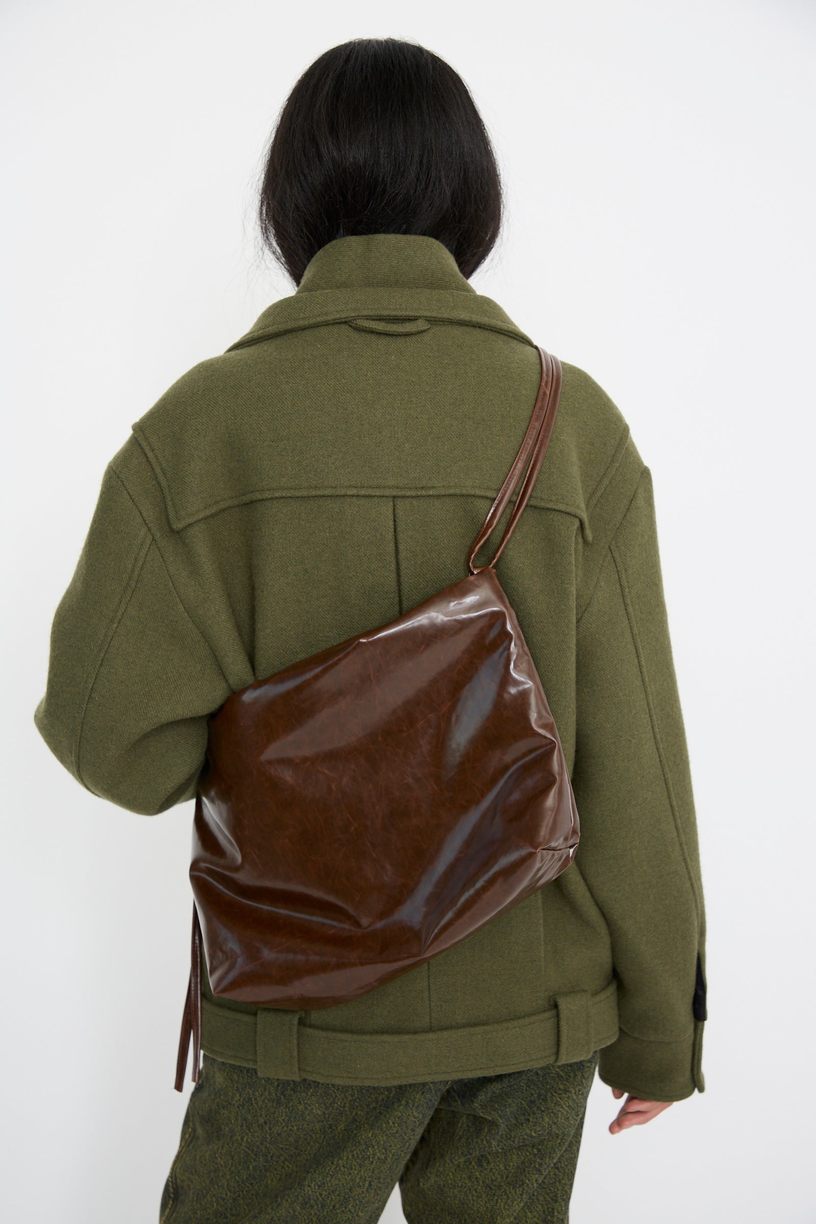 A person wearing a green jacket and dark pants faces away, sporting the Loop Skai Bag in Dark Brown by Kassl, featuring an adjustable strap across their back.