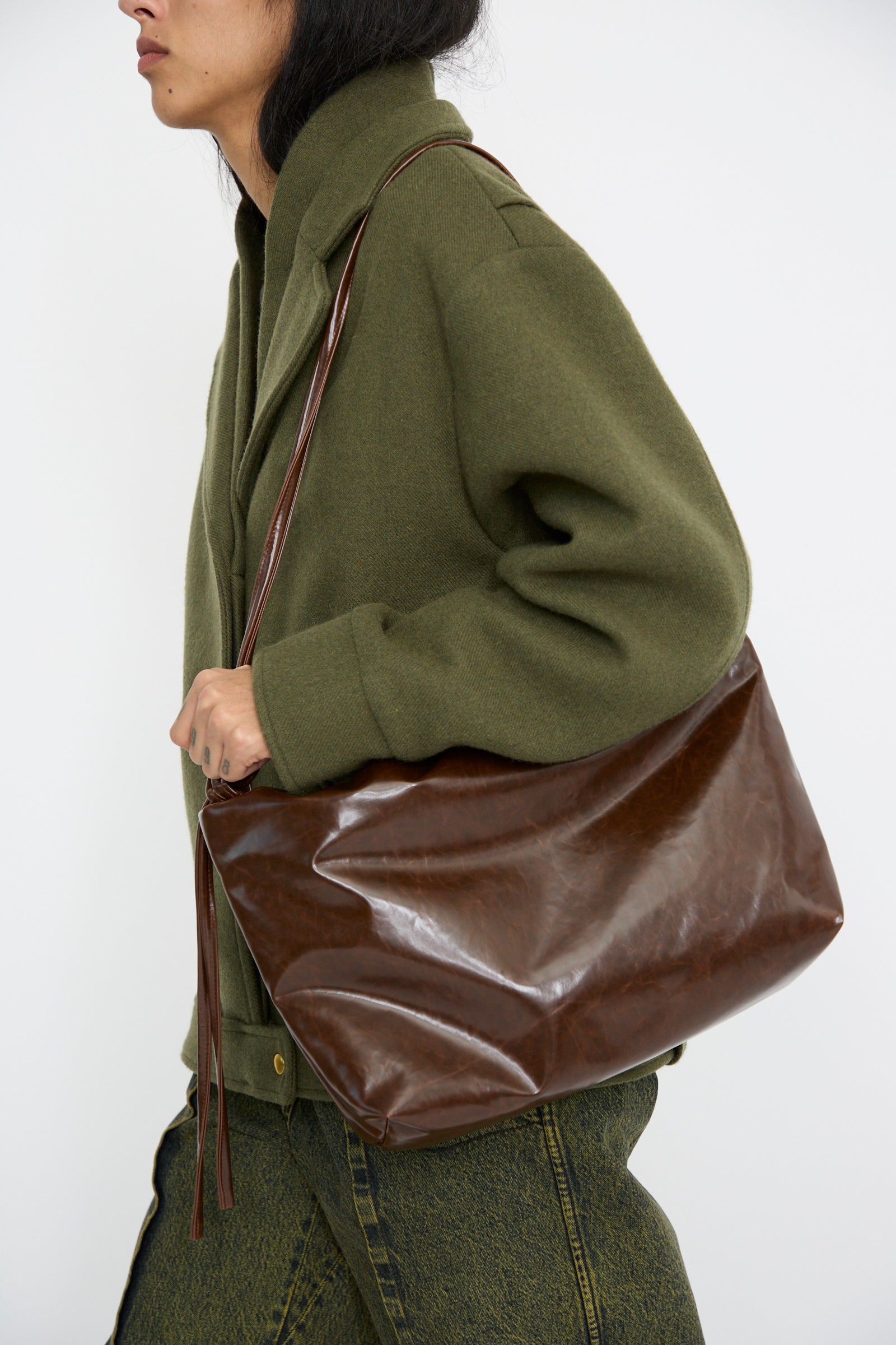 A person in a green jacket carries the Loop Skai Bag in Dark Brown, featuring an adjustable strap, by Kassl over their shoulder against a white background.