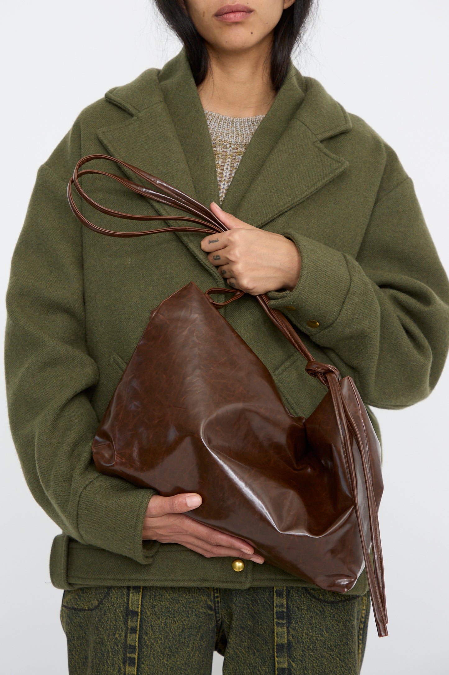 A person wearing a green coat holds the Kassl Loop Skai Bag in Dark Brown, featuring an adjustable strap, against a white background. 