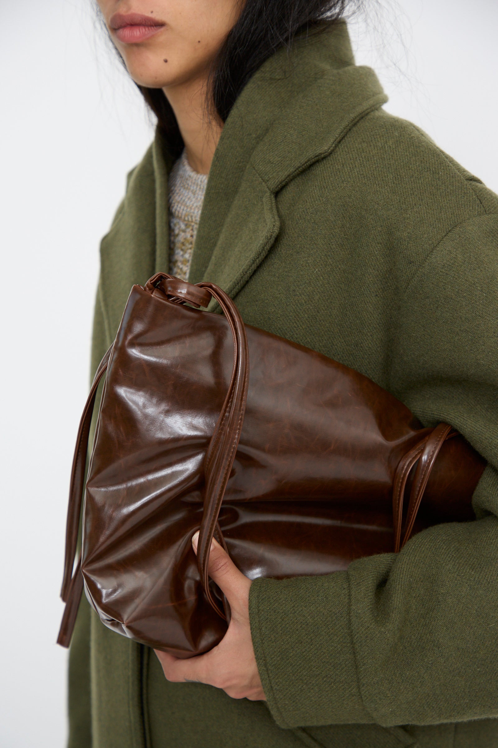 A close up of a  person in a green coat holds the Kassl Loop Skai Bag in Dark Brown, under their arm, against a white background. 