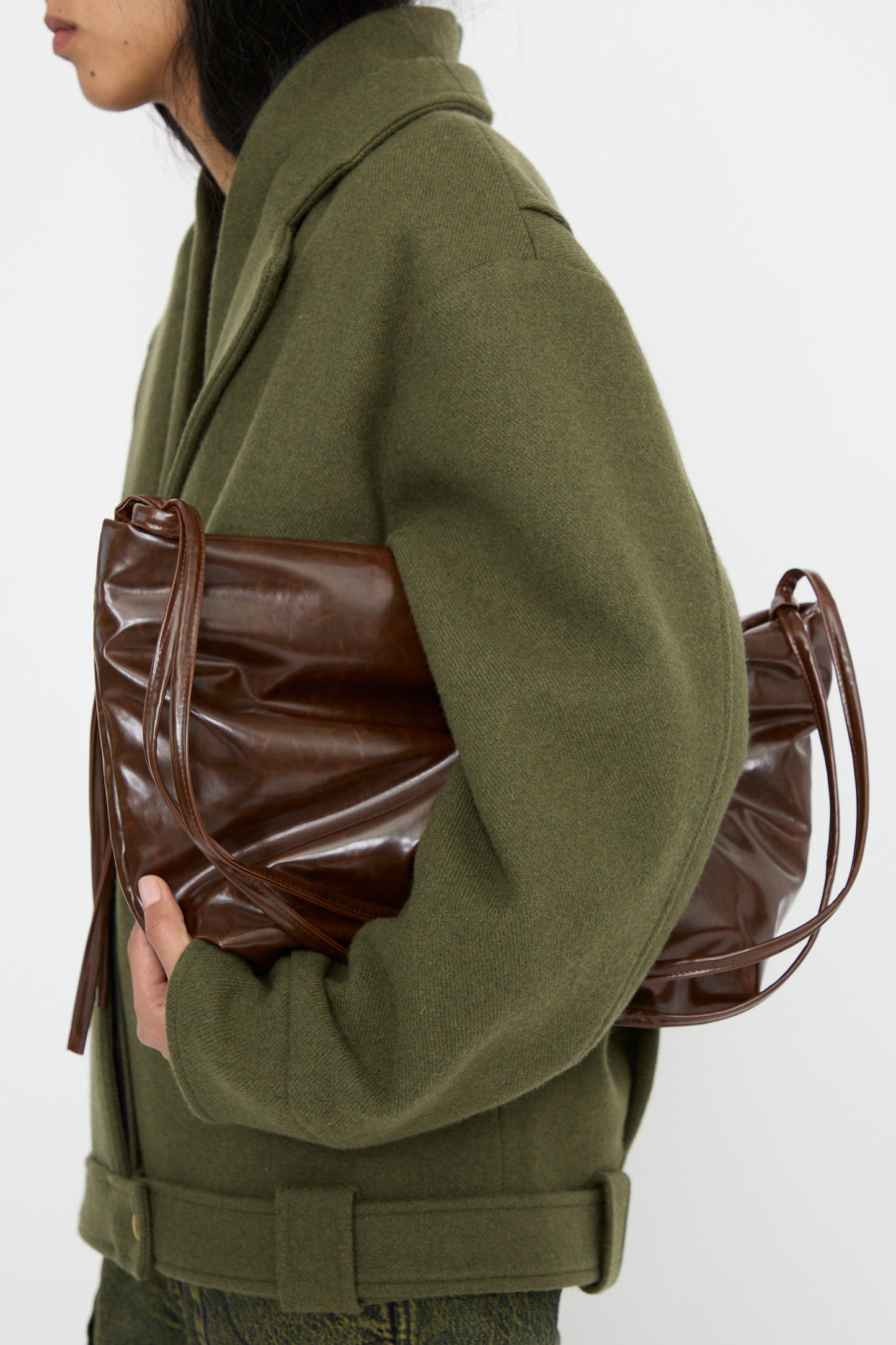 A person in a green coat holds the Loop Skai Bag in Dark Brown by Kassl, with its adjustable strap tucked under their arm, standing against a plain white background.