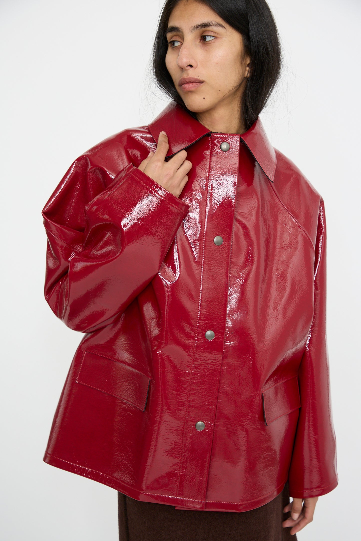 A person is dressed in the Original Hip Lacquer Jacket in Wool Red by Kassl, featuring an eye-catching Fisherman-inspired style with its shiny vinyl finish, large buttons, and front pockets. 