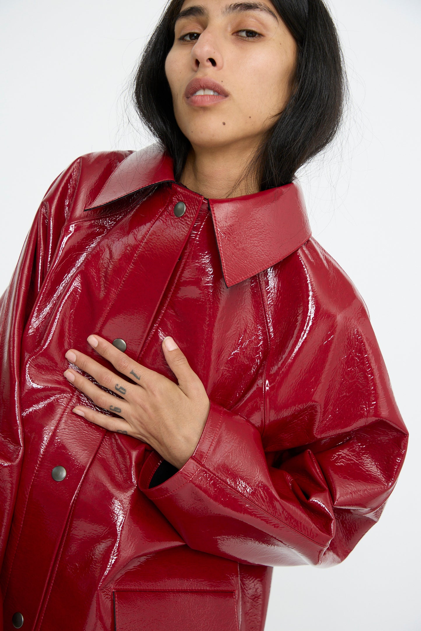 Person in an Original Hip Lacquer Jacket in Wool Red by Kassl, with a shiny finish, pointed collar, and raglan sleeves, posing with one hand on their chest against a plain background.