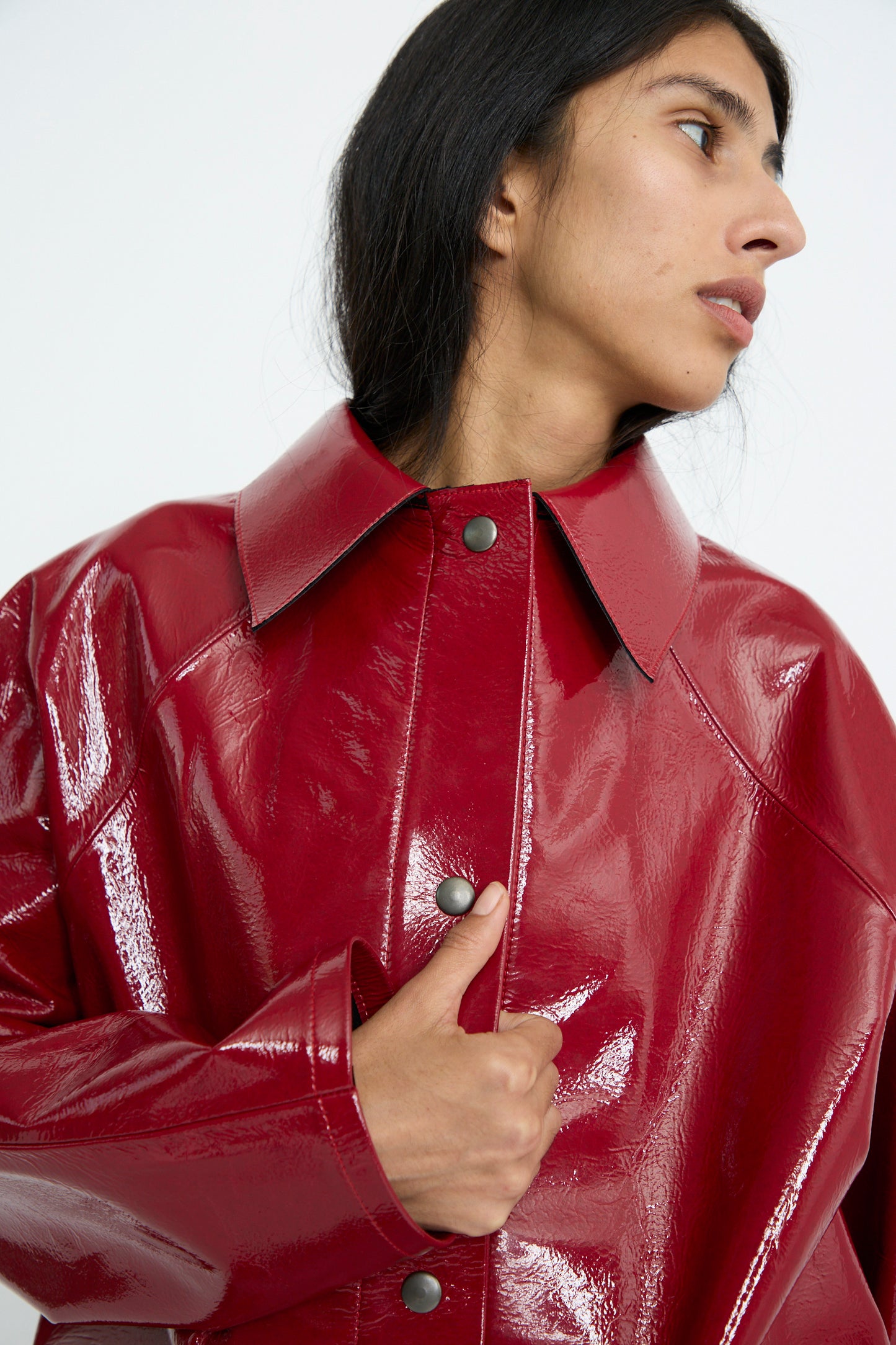 A person wearing the Original Hip Lacquer Jacket in Wool Red by Kassl, featuring button details and raglan sleeves, gazing to the side against a white background. 