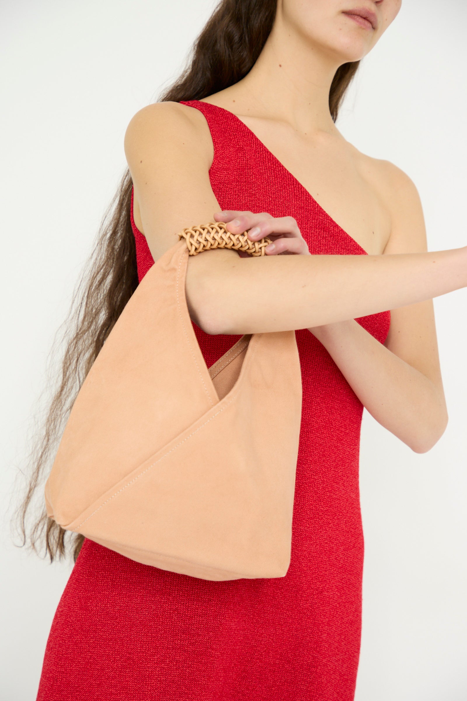 A woman in a red one-shoulder dress elegantly holds Kamaro'an's Woven Triangle Bag 38 in Pink and Natural, featuring a woven canvas design and a braided handle.