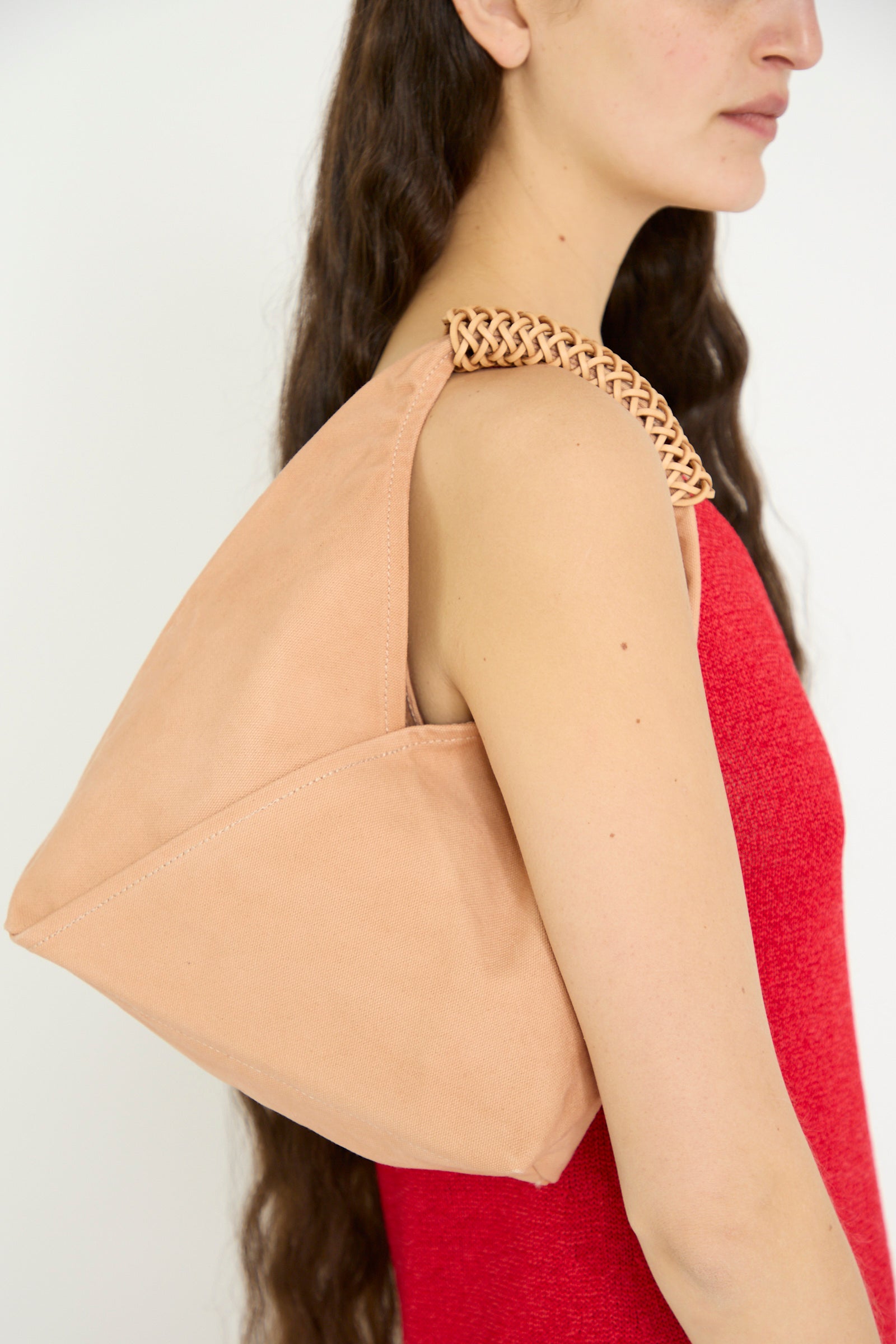 A woman in a red outfit carries the Kamaro'an Woven Triangle Bag 38 in Pink and Natural over her shoulder, against a white backdrop. 