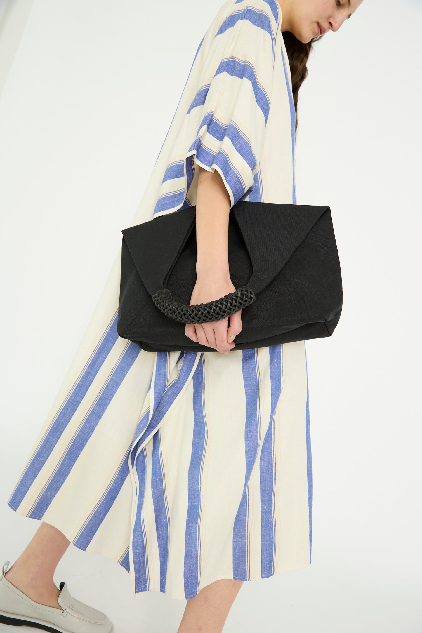 A person in a striped dress walks to the right and holds a large Kamaro'an Woven Triangle Bag 58 in Black with a leather handle.