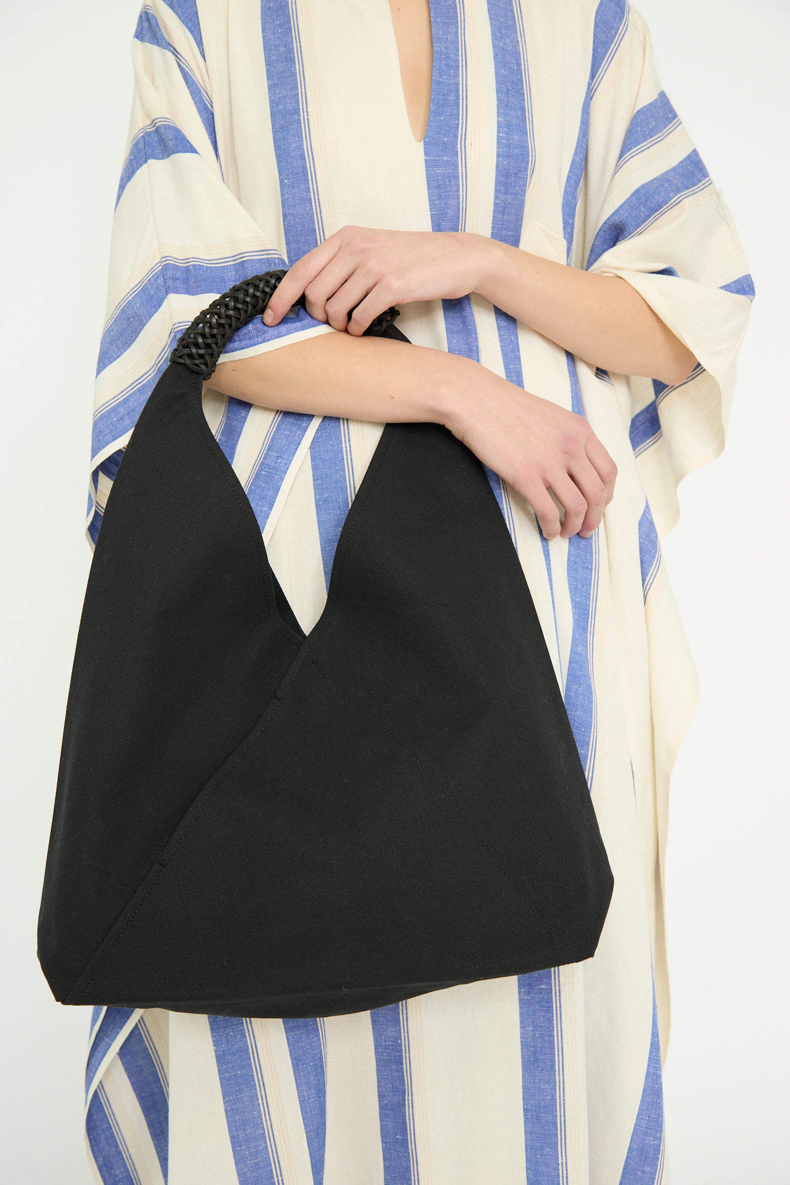 A model in a striped dress carries the Kamaro'an Woven Triangle Bag 58 in Black, against a white backdrop. 