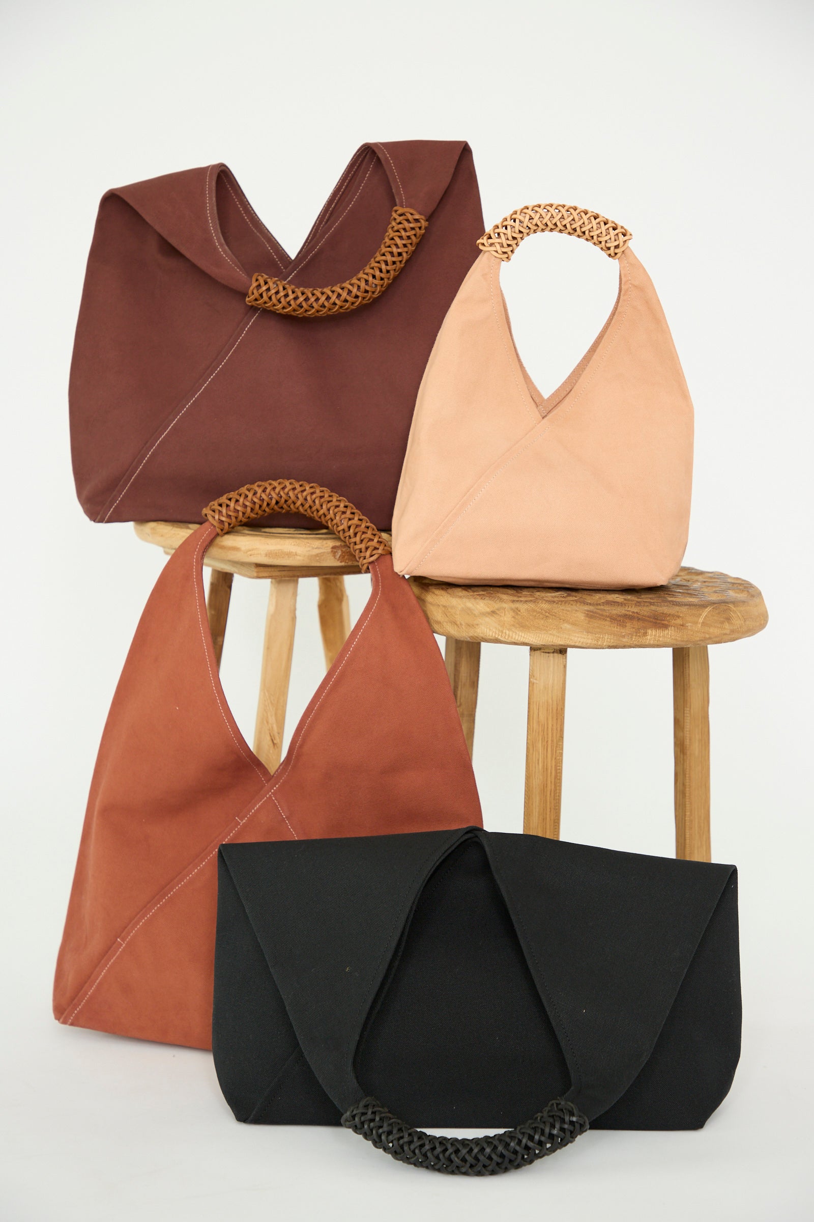 Four Kamaro'an Woven Triangle Bags 58, featuring leather handles in brown, beige, terracotta, and black, are arranged on and around two wooden stools against a white background.