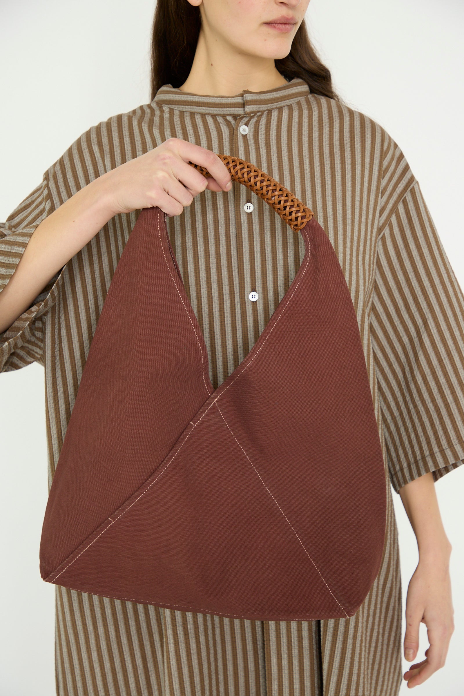 A person carries the Kamaro'an Woven Triangle Bag 58 in Dawn, a brown fabric handbag with a woven handle, against a brown striped dress, in front of a white backdrop. 