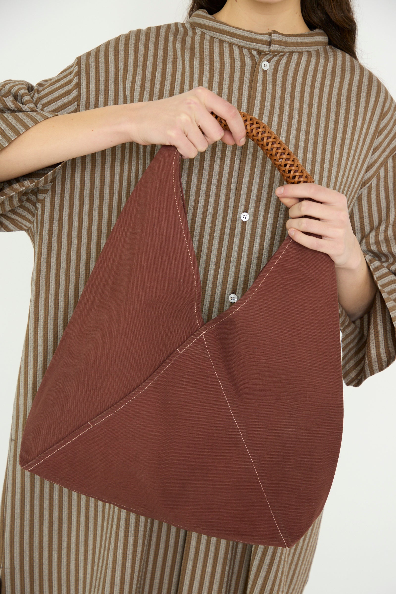 A model carries Kamaro'an's Woven Triangle Bag 58 in Dawn, a purse with a woven canvas handle, held in front of their striped beige and brown outfit.