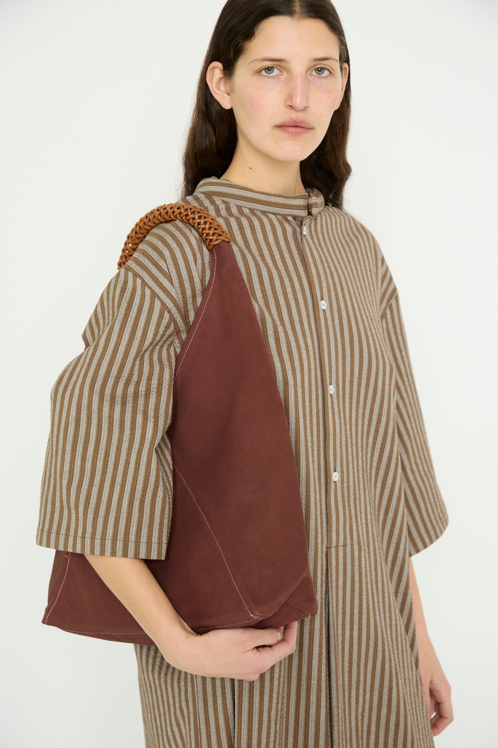 A model wearing a brown striped dress carries the Kamaro'an Woven Triangle Bag 58 in Dawn, complete with woven canvas and leather handles, over their shoulder.