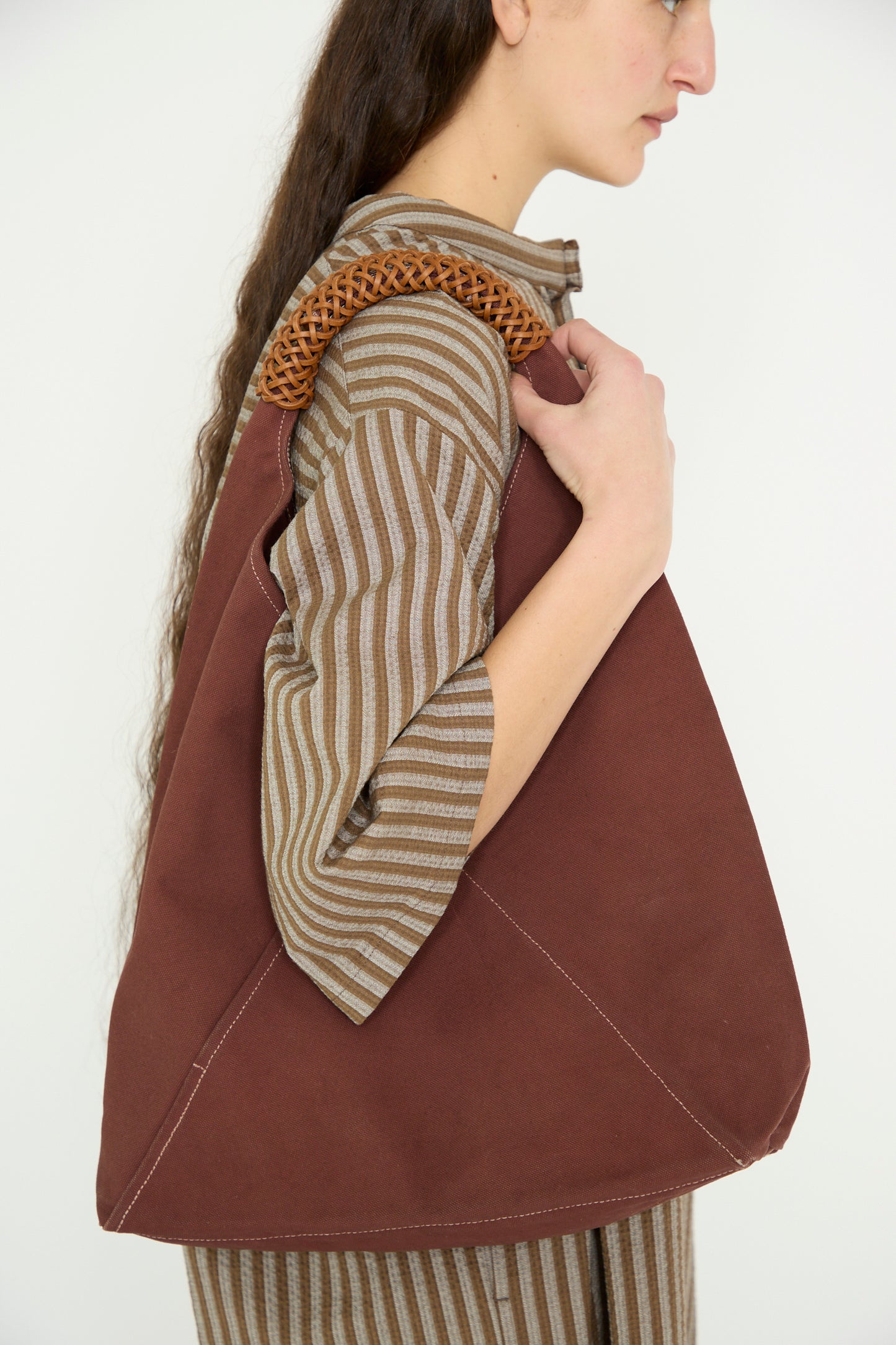 A model facing the side carries a Woven Triangle Bag 58 in Dawn by Kamaro'an on their shoulder, featuring a woven canvas handle, against a white backdrop. 