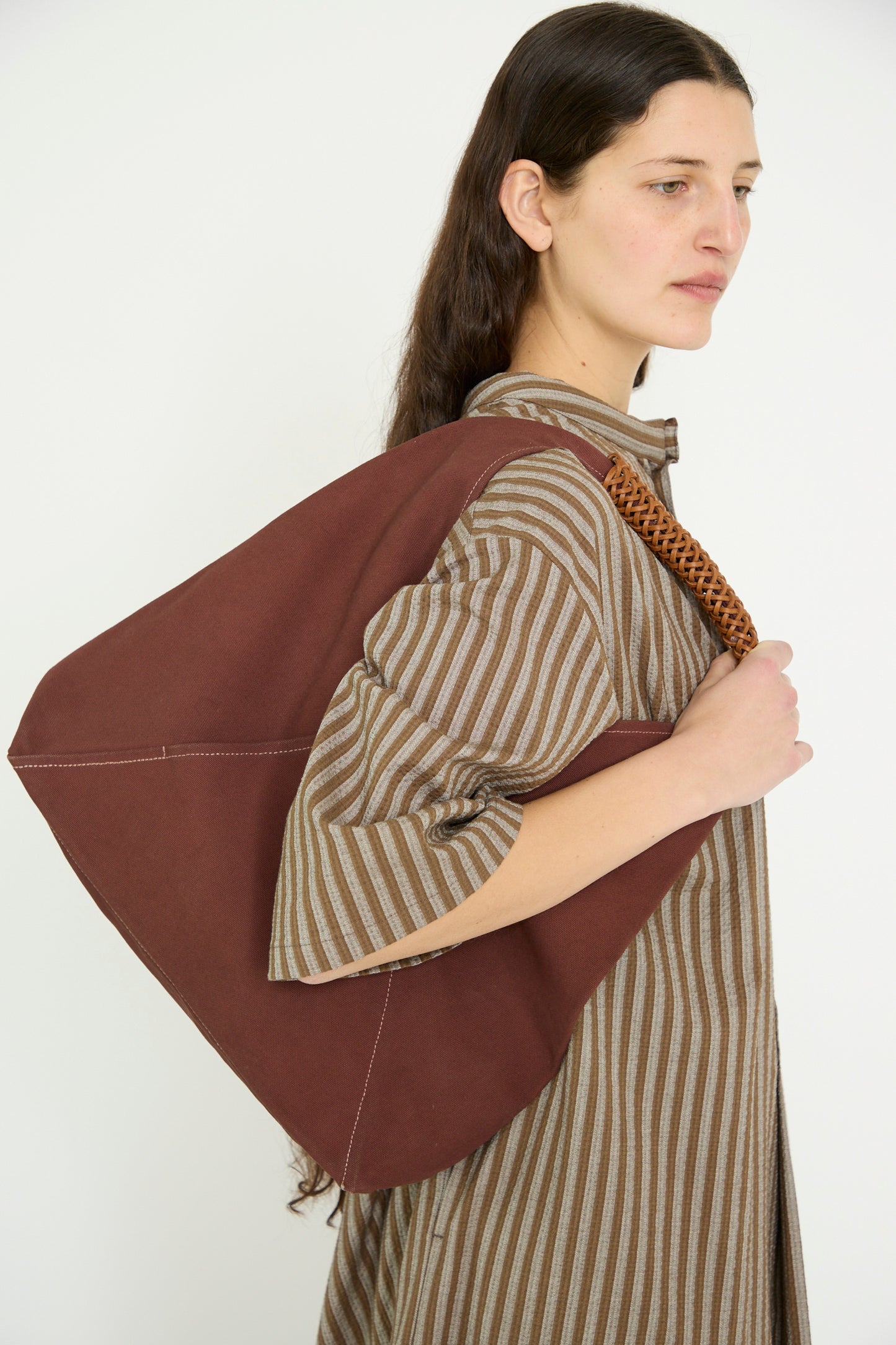 A person with long hair carries a Kamaro'an Woven Triangle Bag 58 in Dawn on their shoulder, wearing a striped dress against a white backdrop. 