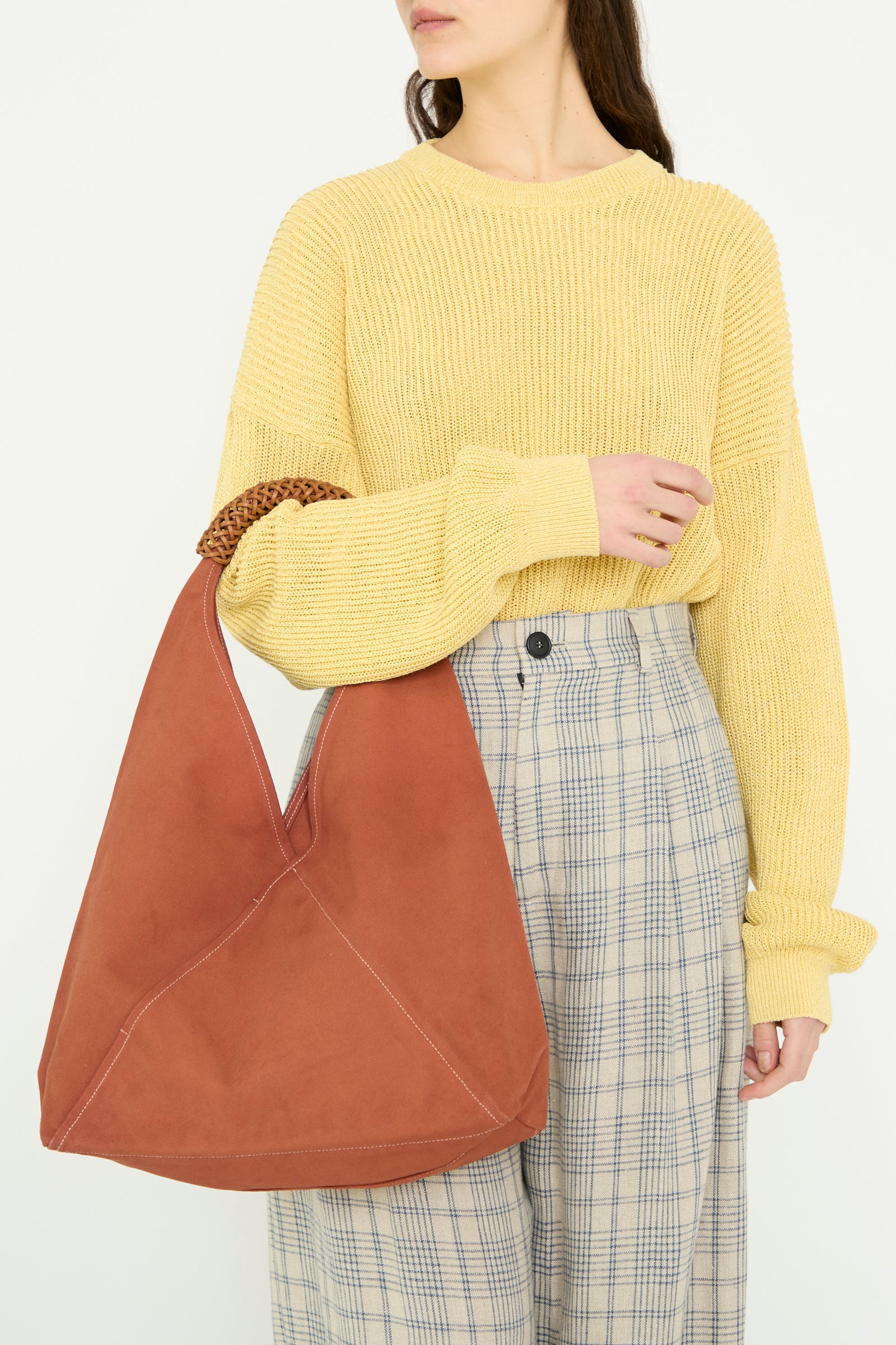 A person wearing a yellow sweater and plaid pants holds the Kamaro'an Woven Triangle Bag in 58 in Ochre Red, featuring a detachable leather handle.