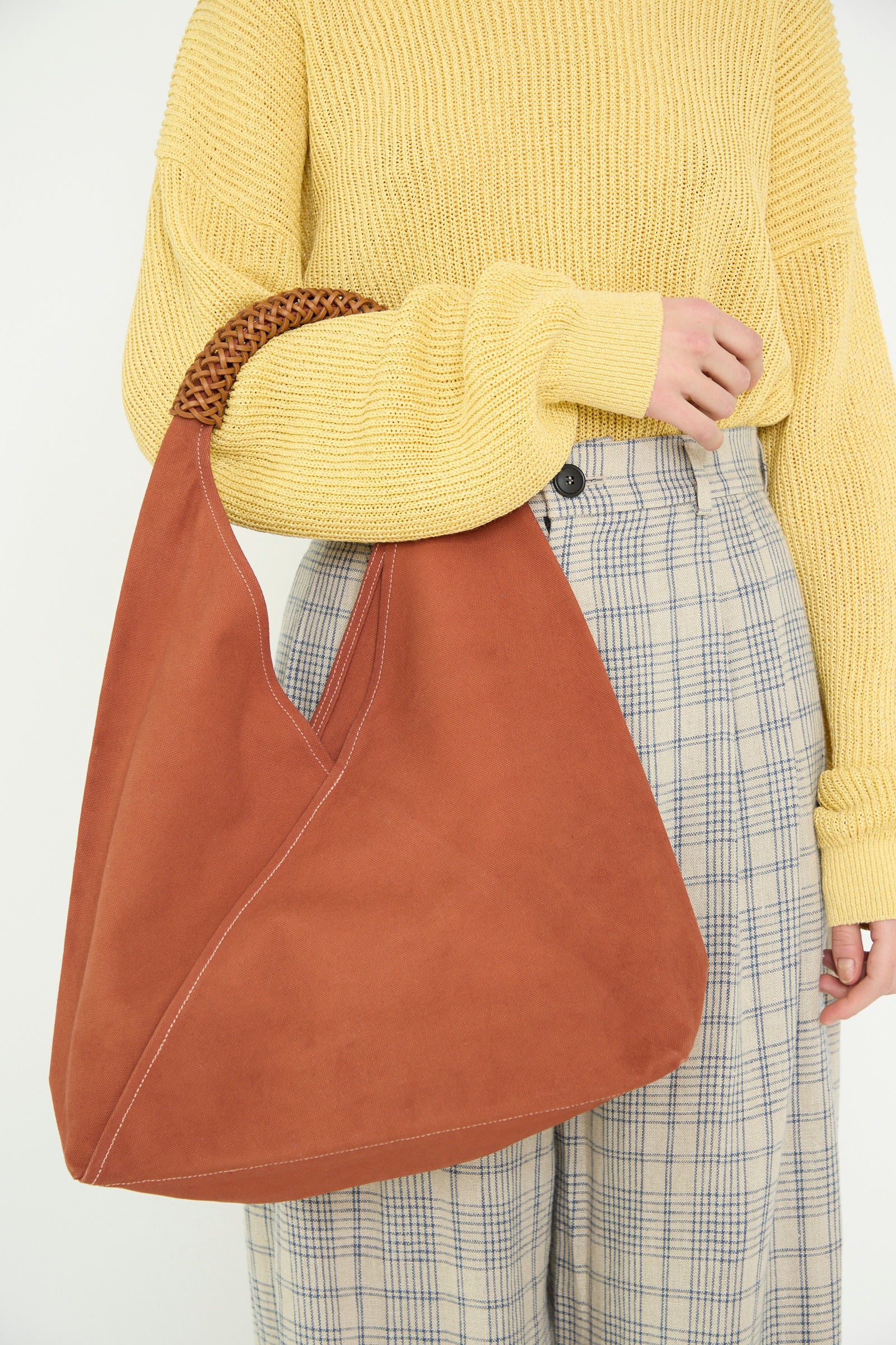 A person in a yellow sweater and plaid pants carries Kamaro'an's Woven Triangle Bag in 58, Ochre Red, seen from below the shoulders. 