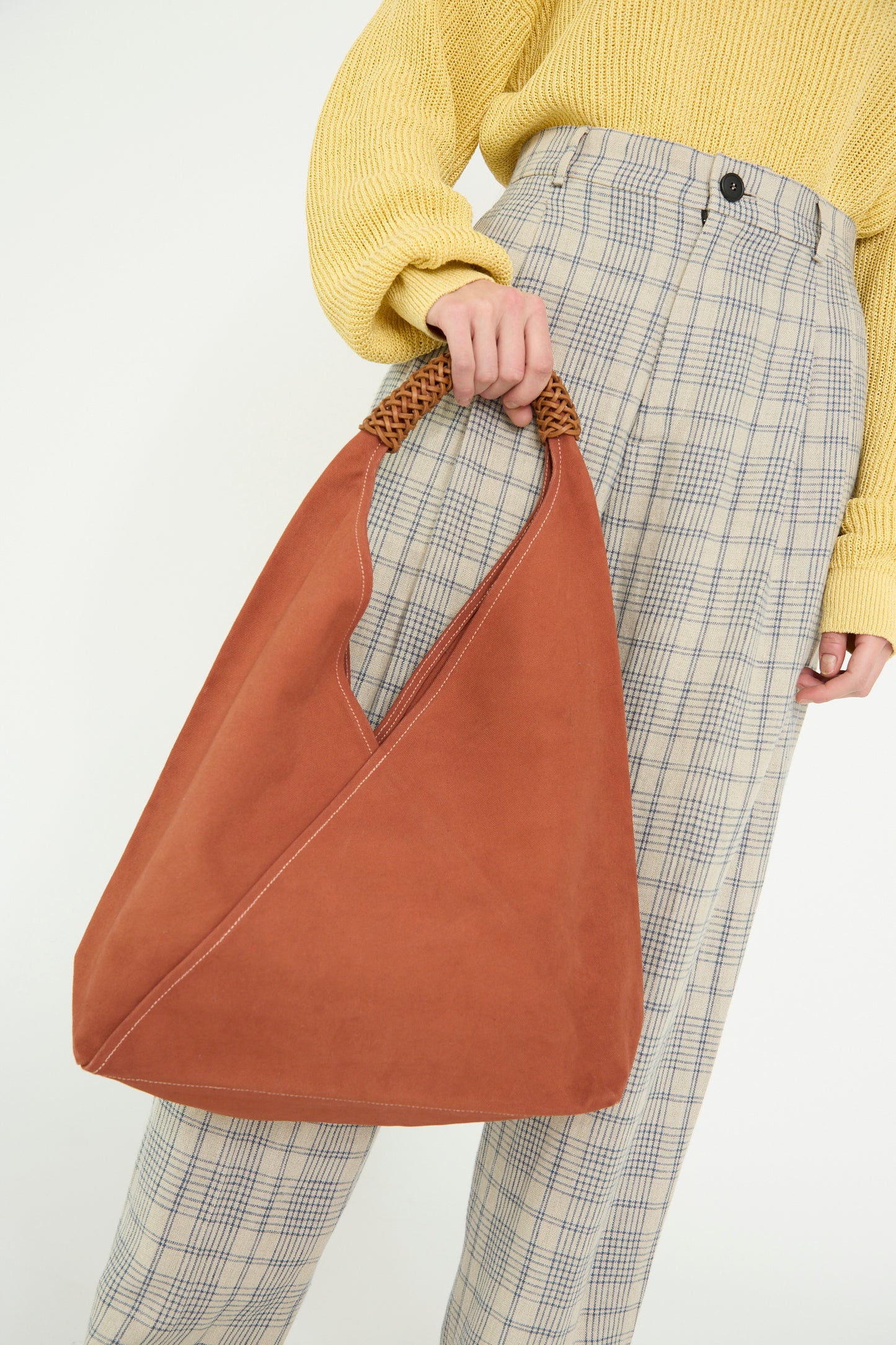 A person holds a large Woven Triangle Bag in 58 in Ochre Red by Kamaro'an, wearing a yellow sweater and plaid pants, against a white backdrop. 