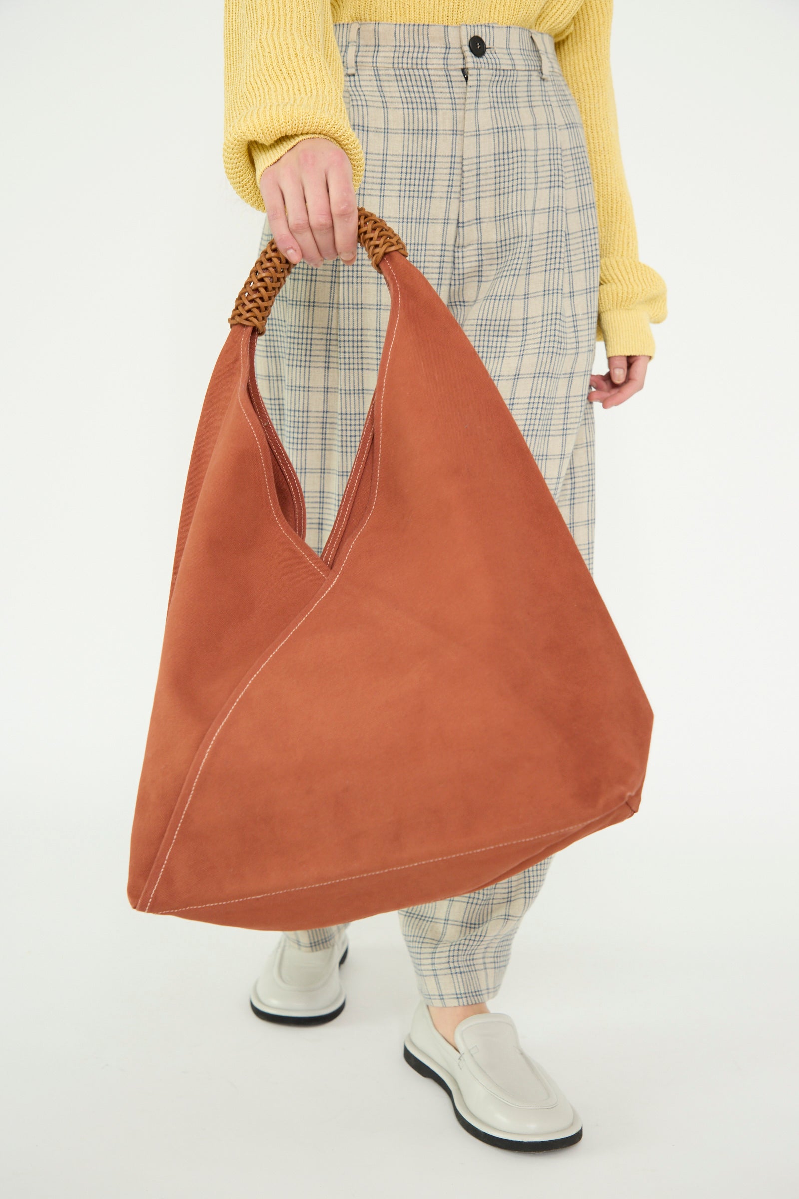 A person holds a Woven Triangle Bag in 58 in Ochre Red by Kamaro'an, wearing a yellow sweater, plaid pants, and white slip-on shoes, posing against a white backdrop. 