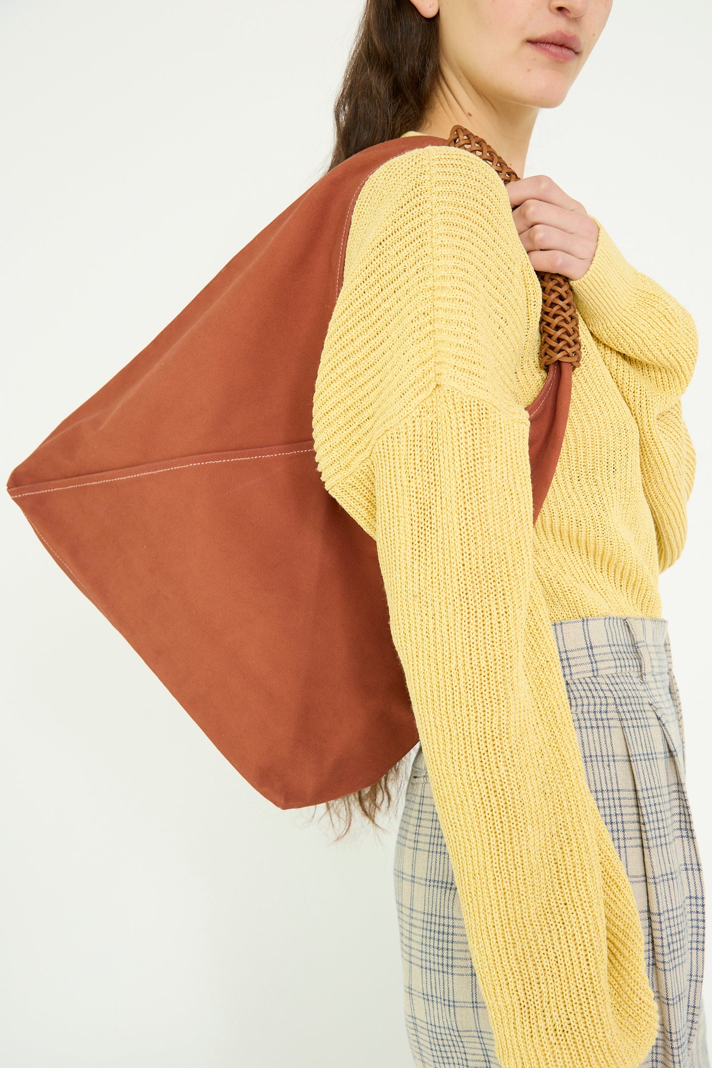 A person in a yellow sweater and plaid pants carries a large ochre red Woven Triangle Bag by Kamaro'an, featuring a leather handle, against a plain backdrop. 