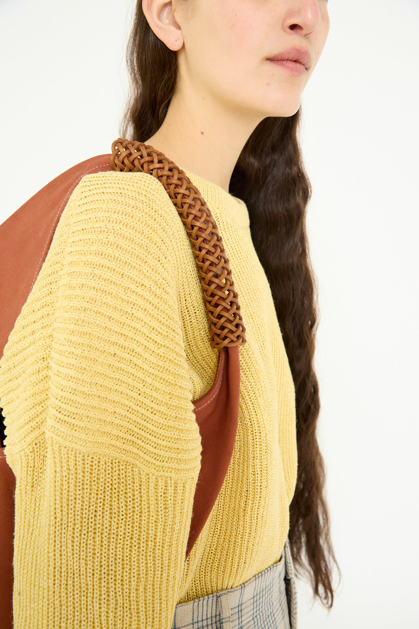 A person with long brown hair wears a yellow sweater, accessorizing their outfit with Kamaro'an's Woven Triangle Bag in 58 in Ochre Red, featuring a brown braided strap.