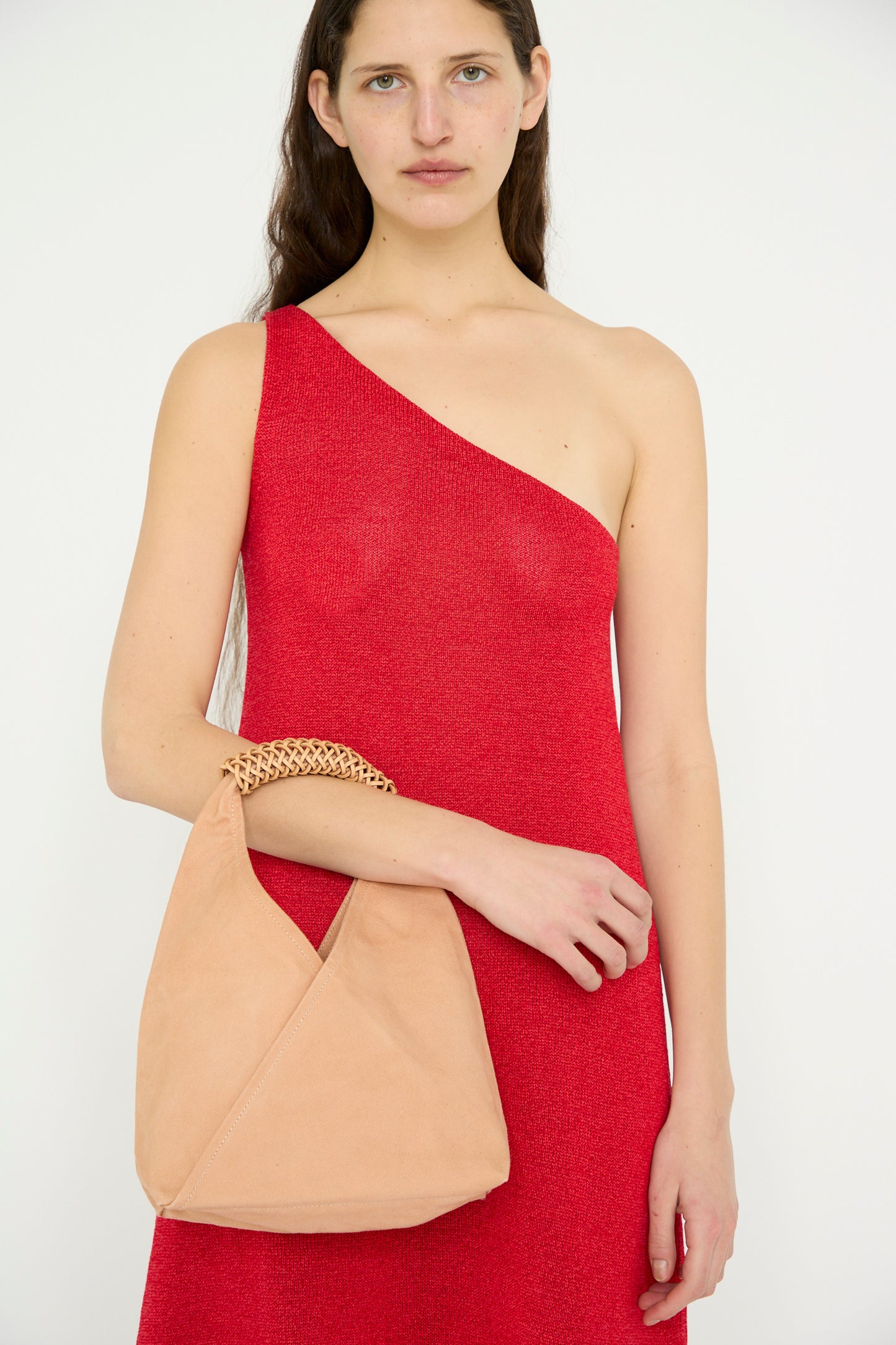 A model in a red one-shoulder dress holds the Kamaro'an Woven Triangle Bag 38 in Pink and Natural, featuring a woven canvas shoulder strap.