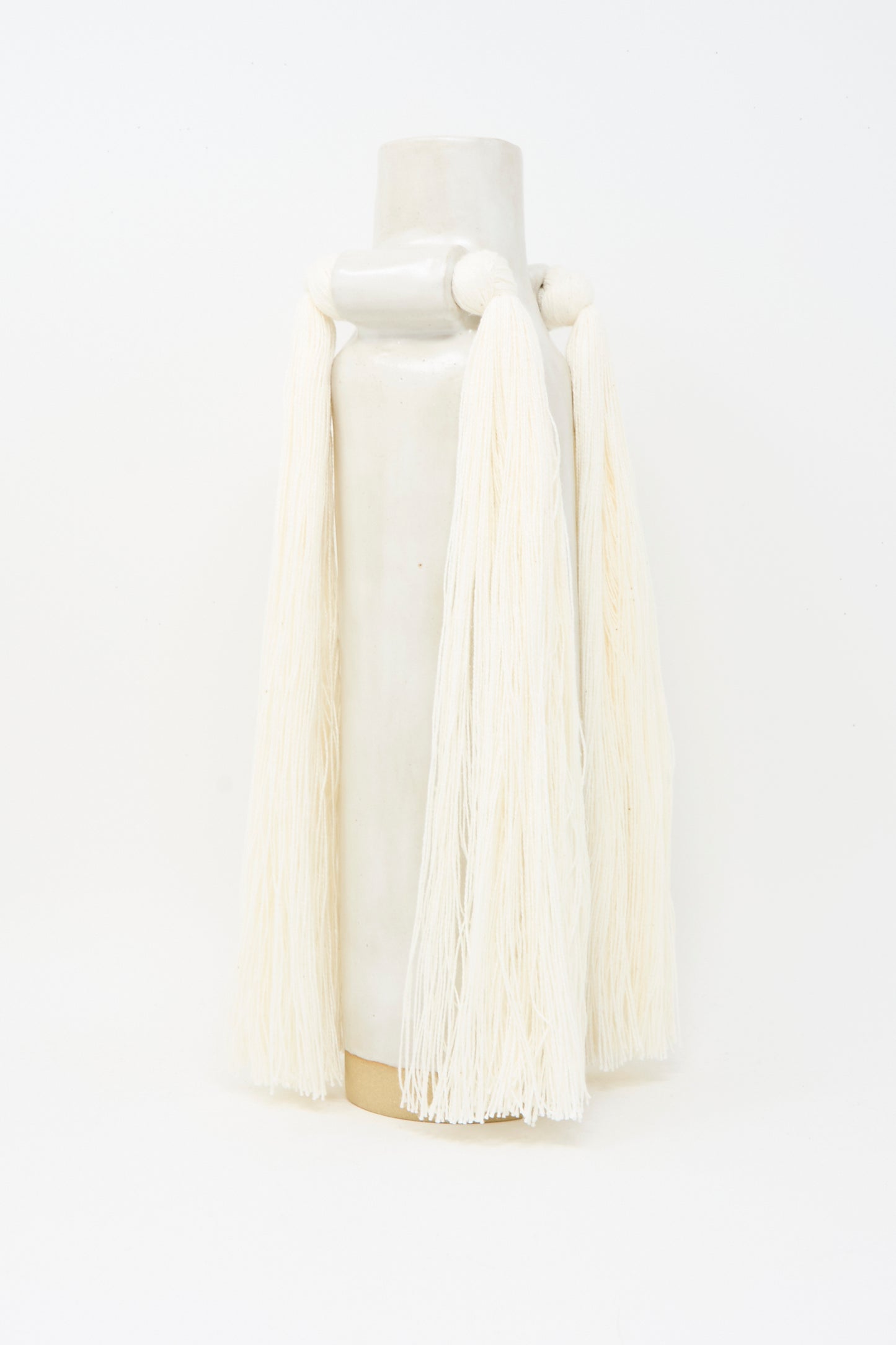 Vase #703 in White by Karen Tinney, a tall handmade stoneware vase with long, thick off-white yarn fringe draped from its sides, is shown against a plain white background.