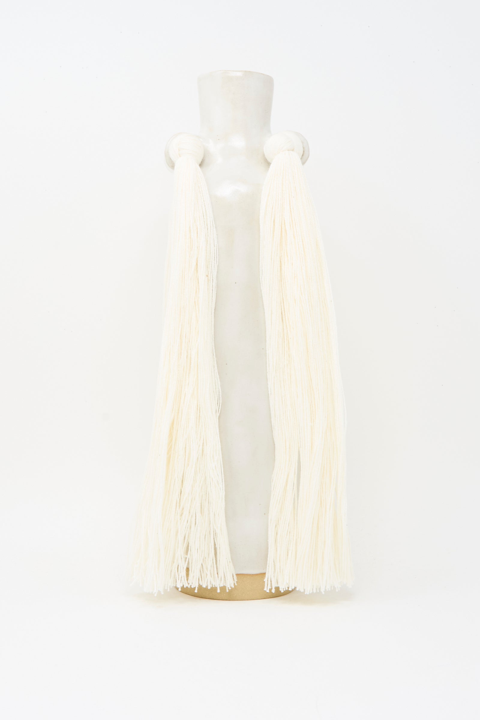 Vase #703 in White by Karen Tinney is a tall, handmade stoneware piece featuring two side handles adorned with long off-white yarn strands. Crafted in Vermont, it stands out against a plain white background.