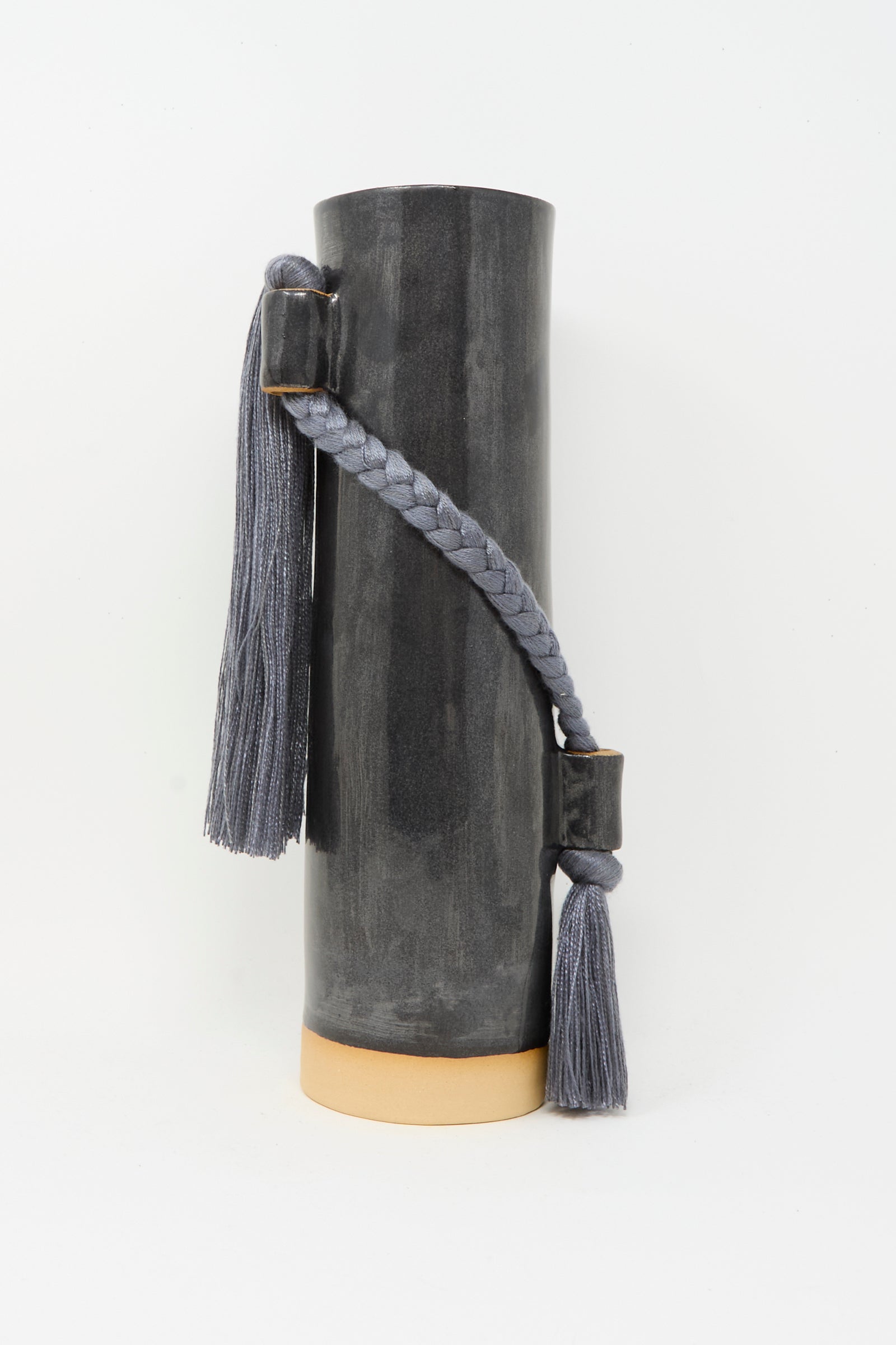 The Karen Tinney Vase #695 in Black is a tall, cylindrical stoneware piece with a light beige base and two gray braided tassels attached to the side with rectangular holders.