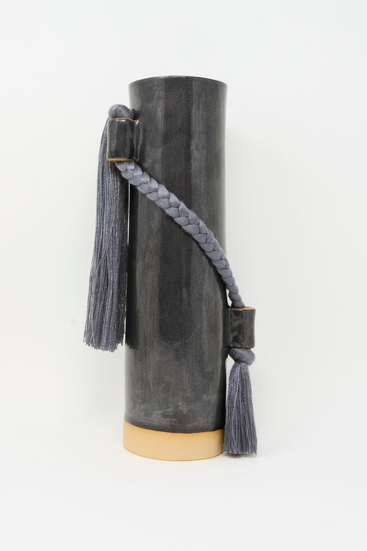 The Karen Tinney Vase #695 in Black is a tall, cylindrical stoneware piece with a light beige base and two gray braided tassels attached to the side with rectangular holders.