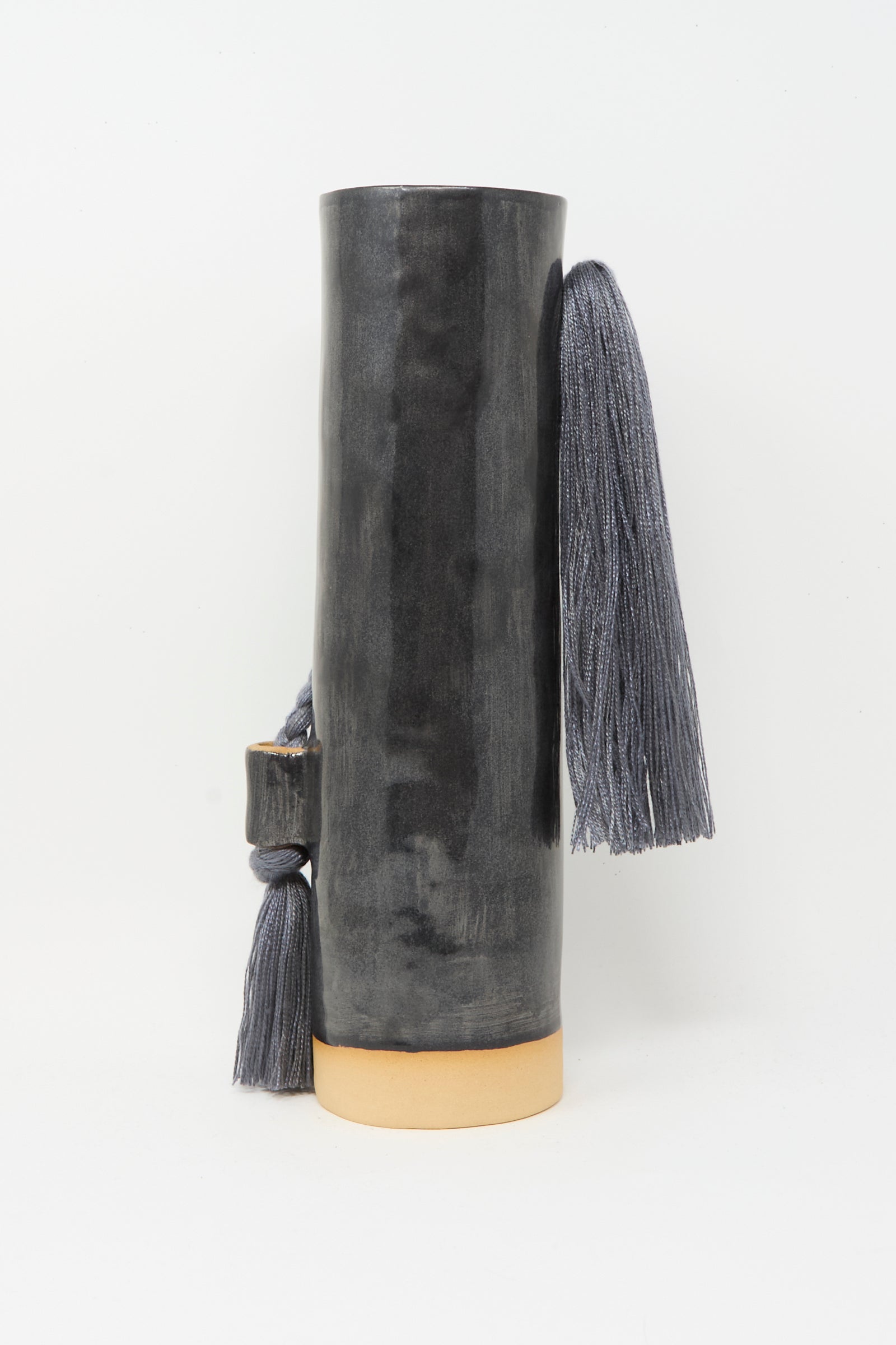 The Karen Tinney Vase #695 in Black is a tall, handmade cylindrical vase with a glossy black stoneware finish, natural beige base, and two sections accented by long gray tassels.