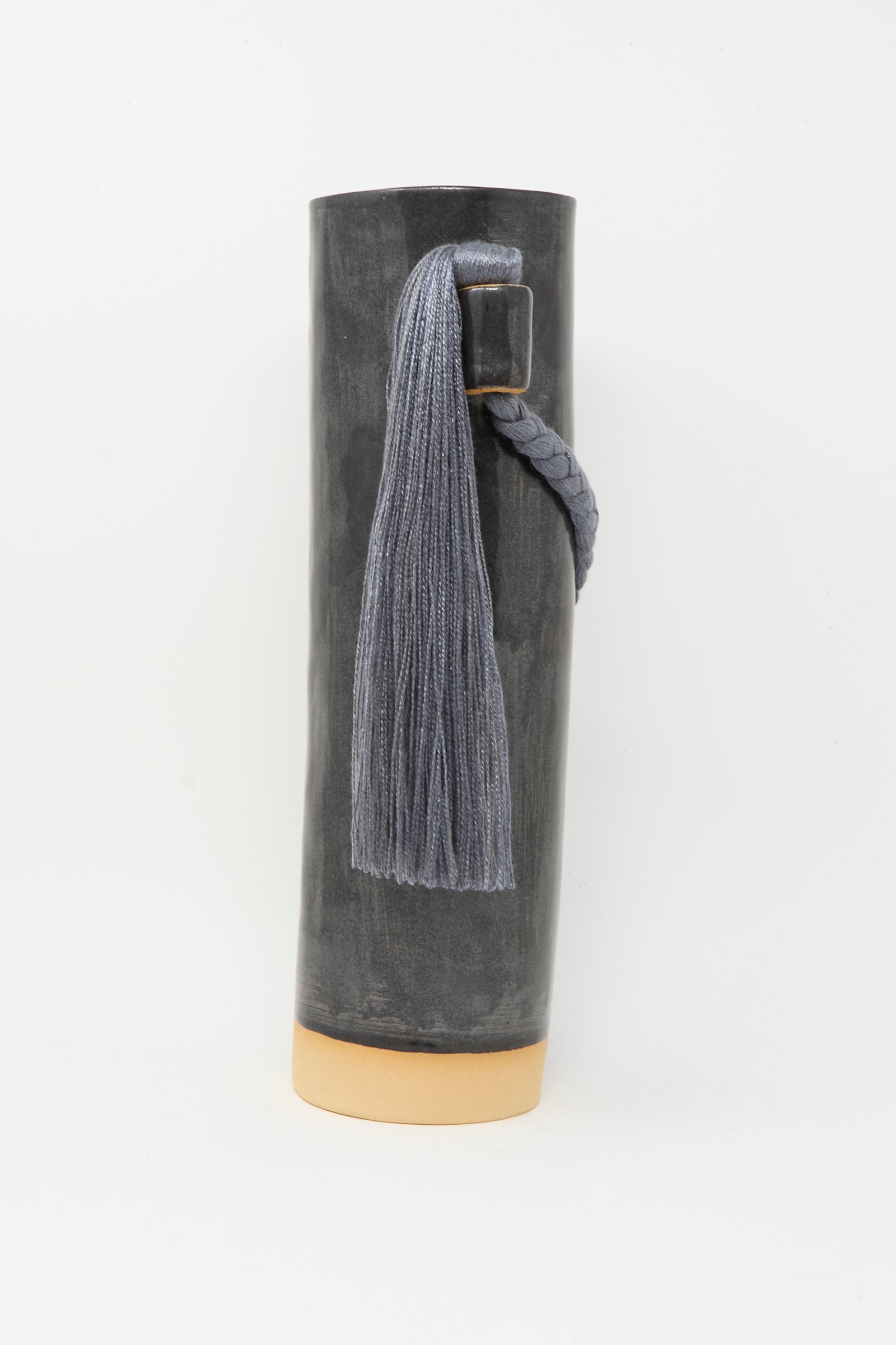 Vase #695 in Black by Karen Tinney is a tall, cylindrical black stoneware vase with a beige base, handmade with a braided rope and gray tassel.