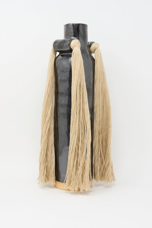 Karen Tinney’s Vase #703 in Black is a tall, glossy stoneware vase featuring two large beige yarn tassels near the top, shown against a plain white background.