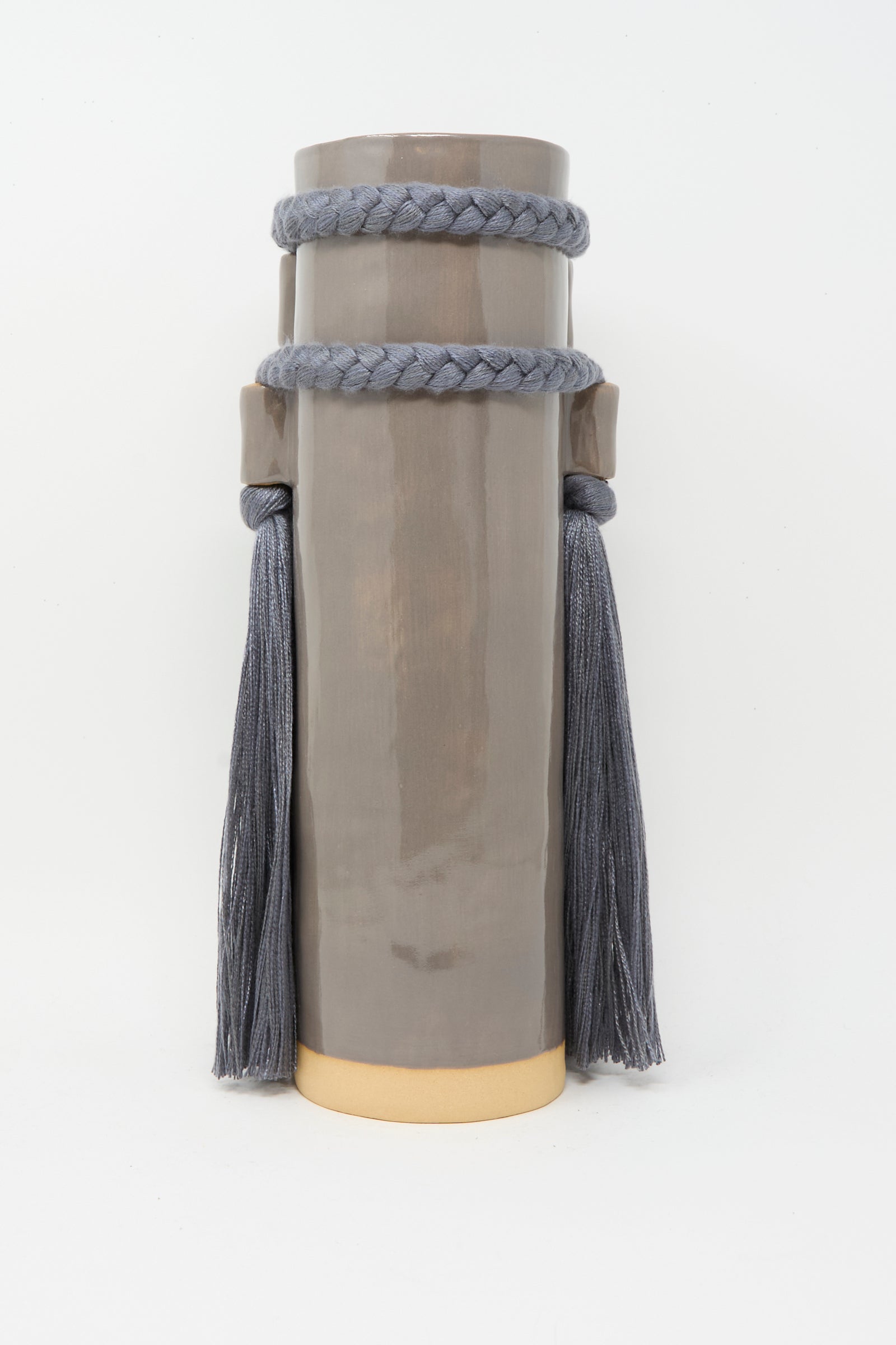 Karen Tinney's Vase #735 in Charcoal is a tall cylindrical handmade stoneware vase featuring a glossy taupe finish, braided rope with long tassels near the top, and a gold-colored base, shown against a white background.