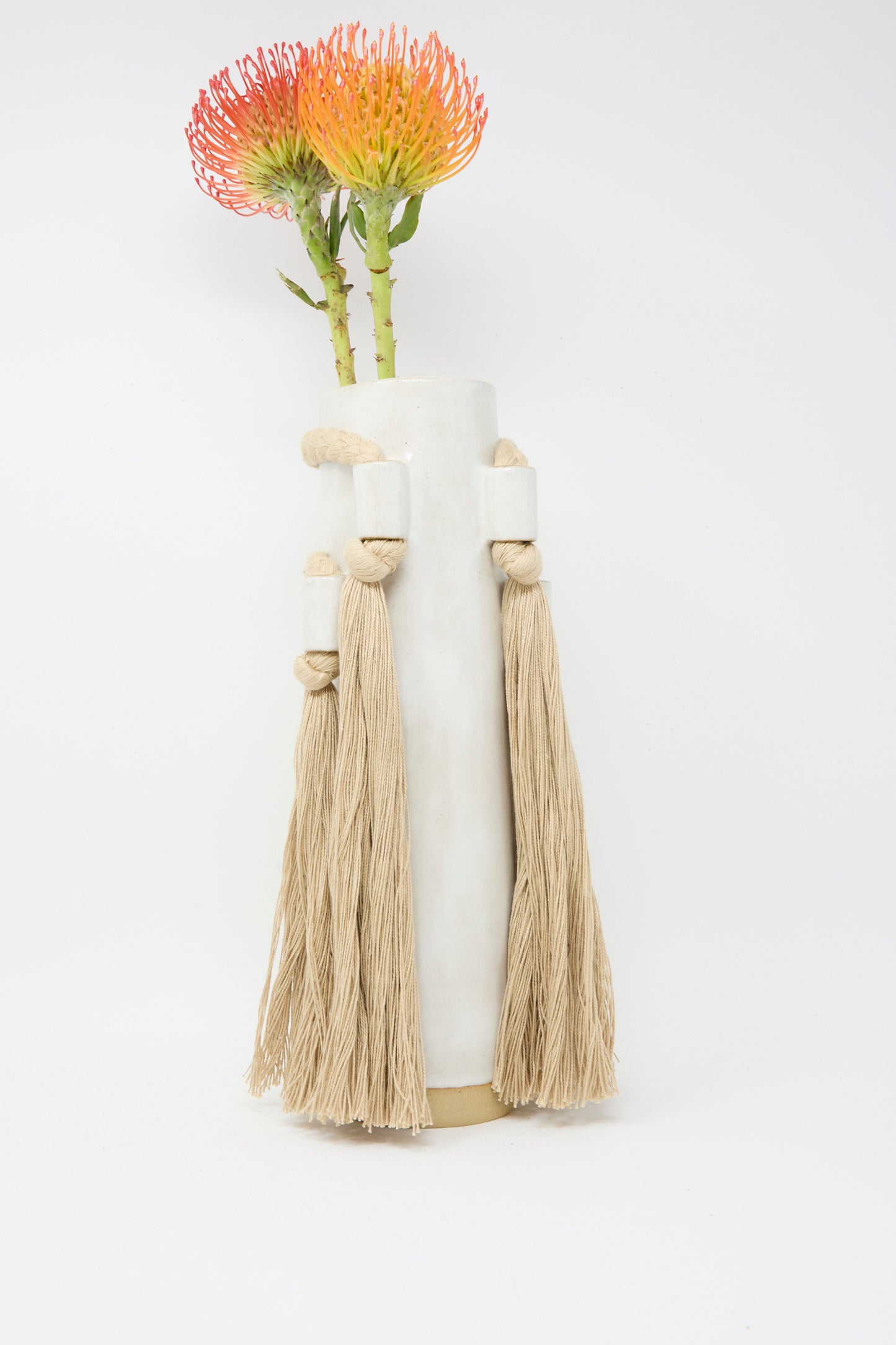 Karen Tinney's Vase #735 in White, a handmade stoneware piece with rope and tassel accents, holds two orange flowers against a plain white background.